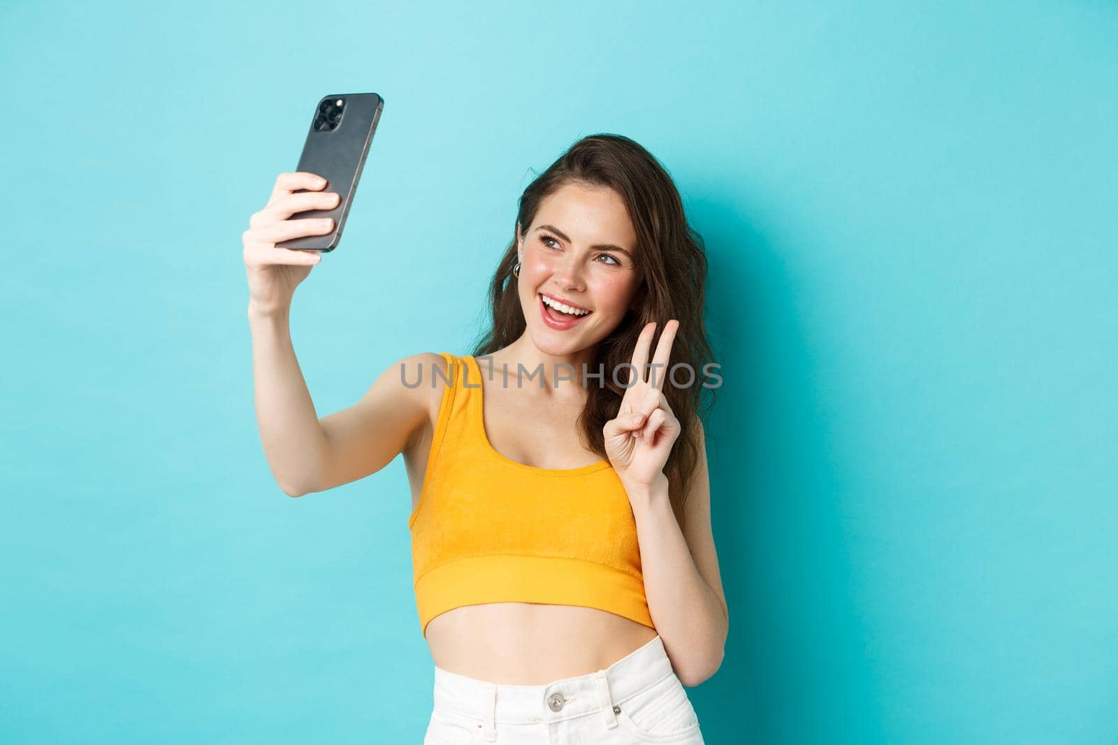Technology and lifestyle concept. Stylish young woman taking selfie in her summer clothes, showing v-sign at smartphone camera, standing over blue background.