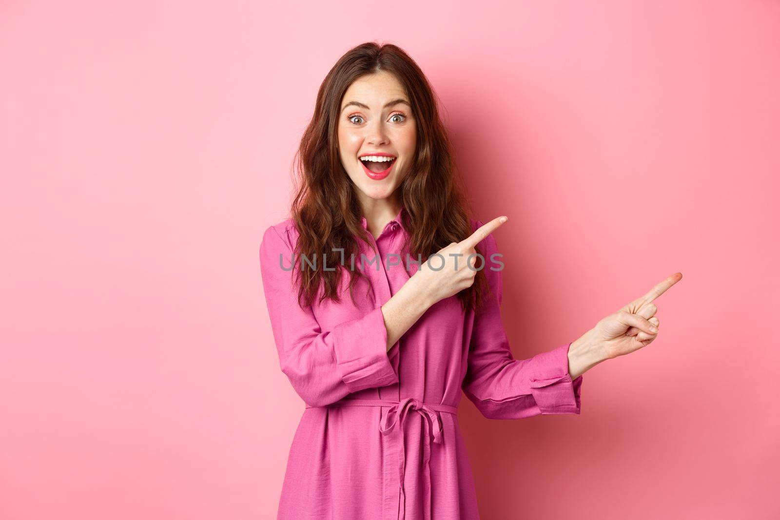 Beauty and fashion. Amazed stylish modern woman pointing fingers right, showing promo on copyspace aside, standing against pink background.