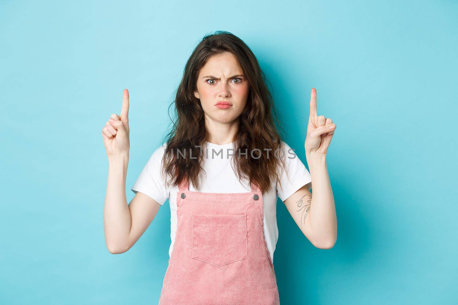 Confused and shocked young woman frowning, staring at camera displeased, demand answers, pointing fingers up on top banner, standing over blue background.