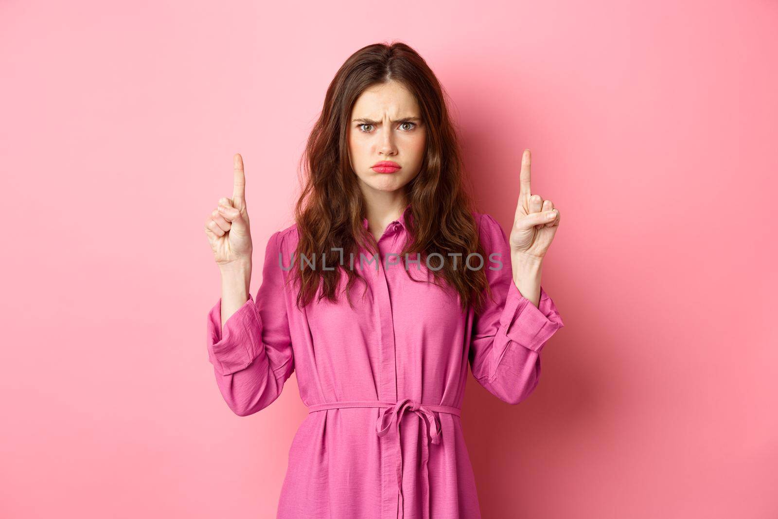 Disappointed glamour girl complaining, sulking with coy expression, pointing fingers up, show something unfair, standing jealous against pink background.