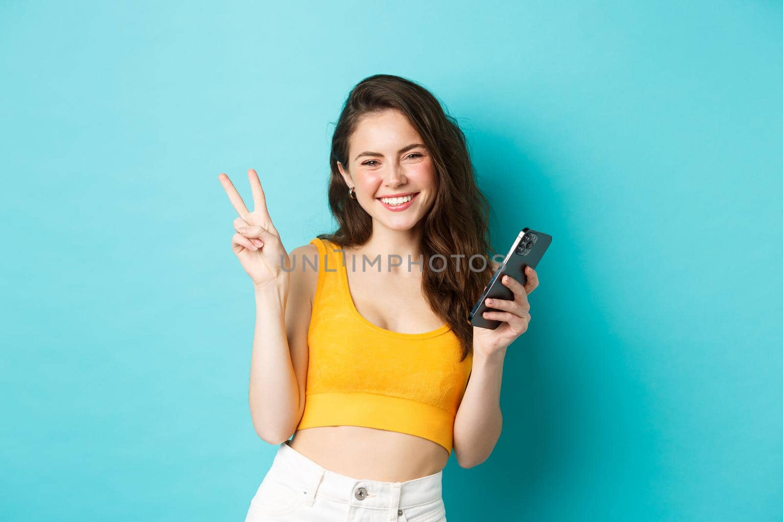 Technology and lifestyle concept. Beautiful cheerful girl sending positive vibes, smiling and showing peace sign, using cellphone, standing against blue background.