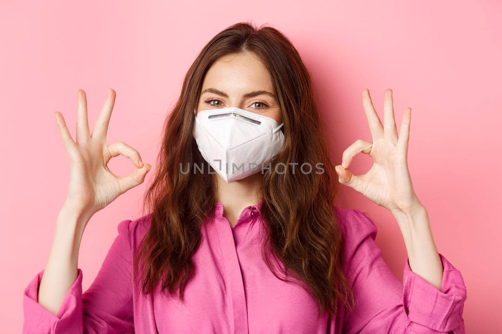 Coronavirus, preventive measures and health concept. Close up portrait of good-looking woman wears her medical respirator from covid-19, shows okay signs and smiling pleased.