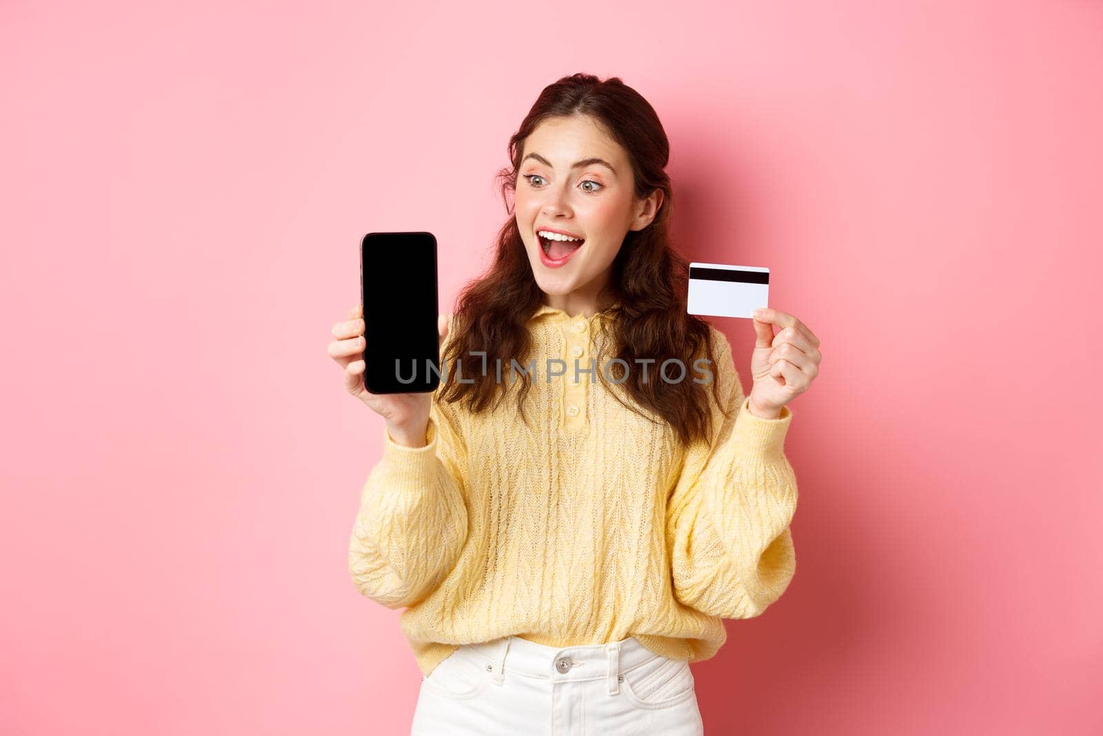 Technology and online shopping. Excited attractive girl showing smartphone screen, plastic credit card, look amazed at phone, standing against pink background by Benzoix