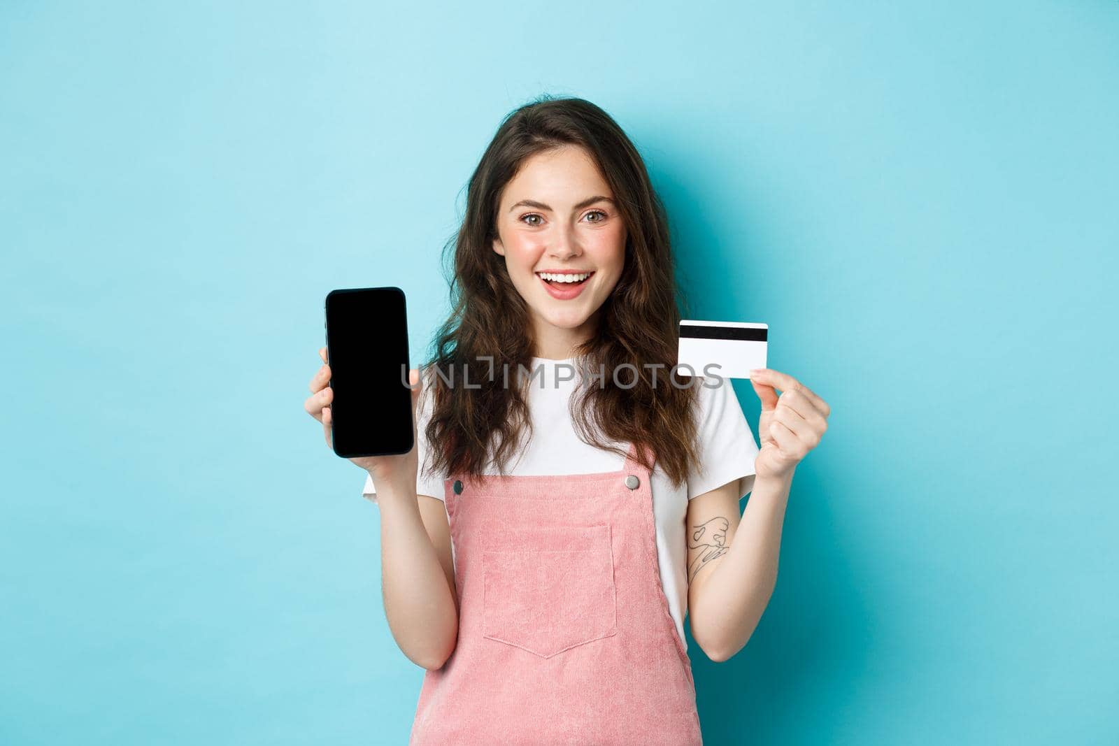 Image of beautiful smiling woman in stylish outfit, showing plastic credit card and empty smartphone screen, recommend app, standing over blue background by Benzoix