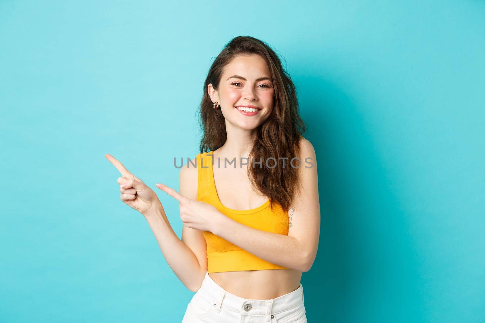 Beautiful young woman looking happy, smiling with teeth, inviting to check out event promo, pointing fingers left at copy space, standing over blue background.