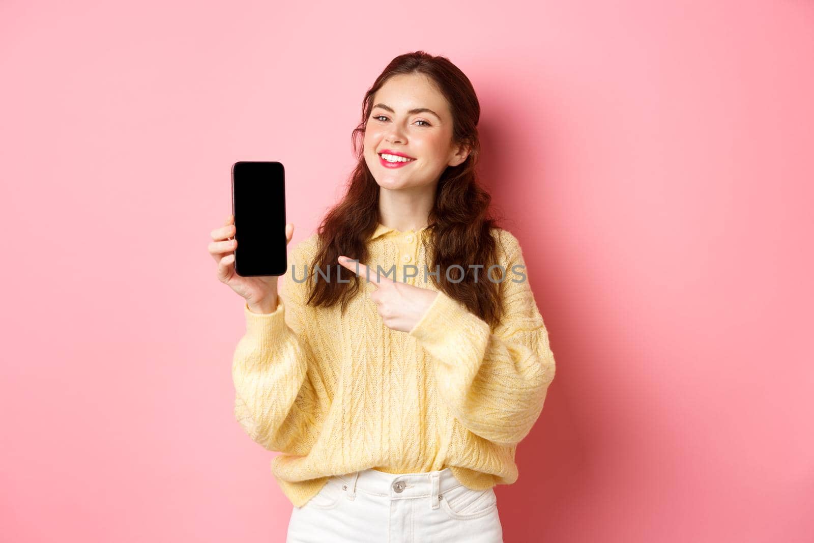 Portrait of attractive young woman demonstrates promo on phone, pointing finger at smartphone and smiling, standing against pink background.