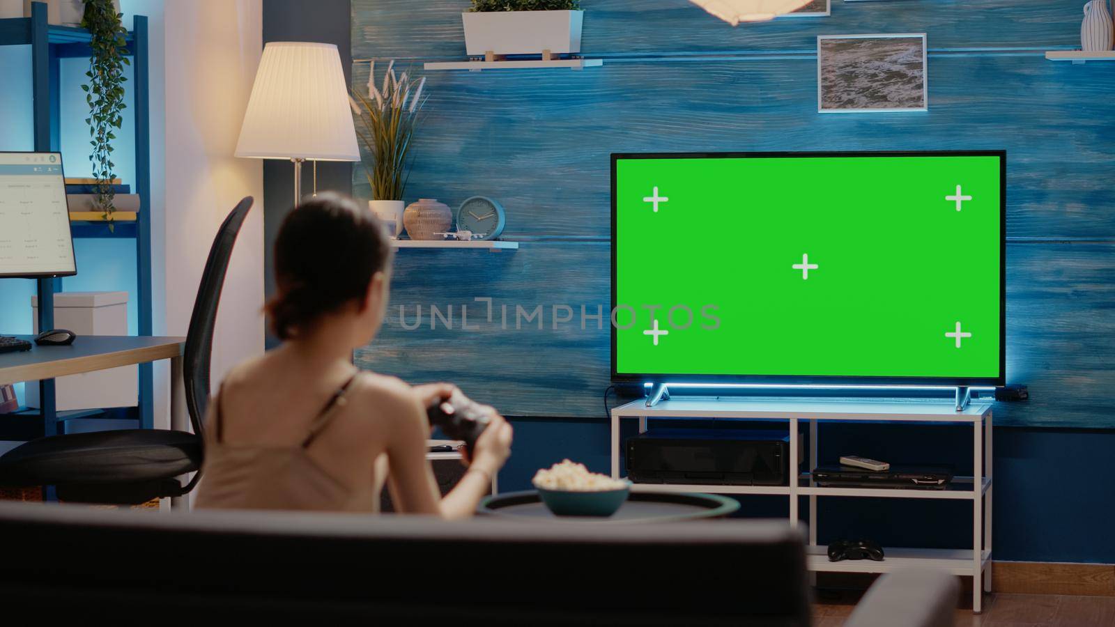 Person using joystick and green screen display on tv sitting in living room on couch. Caucasian woman with chroma key used for blank mockup template and isolated virtual background