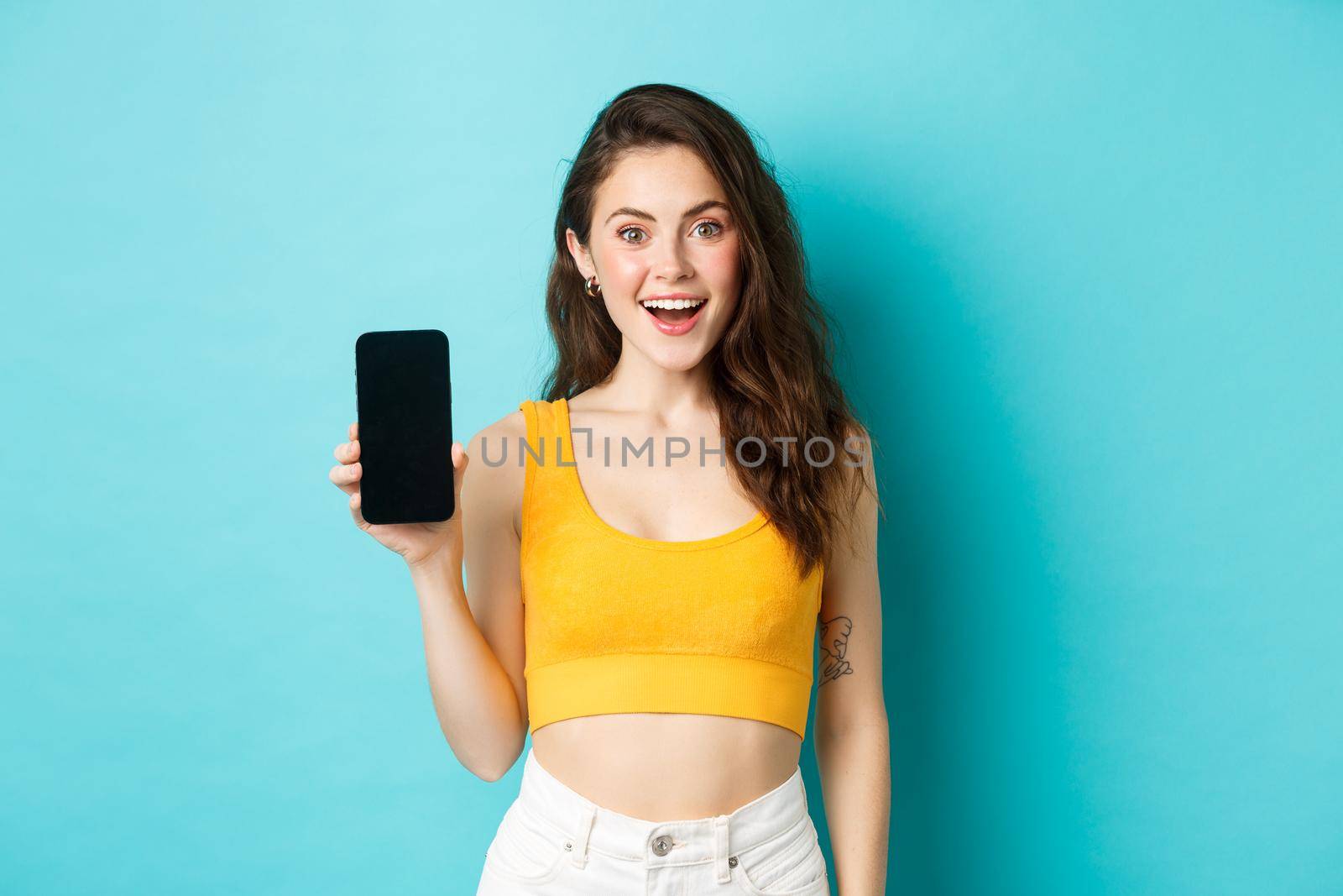Excited young woman in yellow crop top showing blank smartphone screen, demonstrate your advertisement on display, standing over blue background.