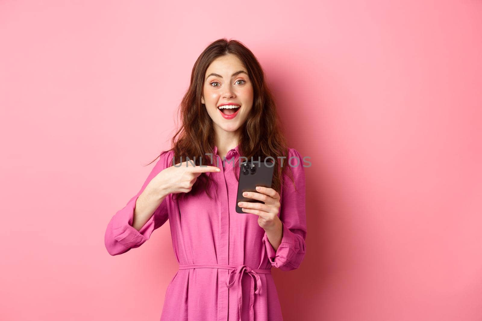 Young woman pointing finger at her phone and smiling, talking about smartphone app, showing something interesting online, standing over pink background.