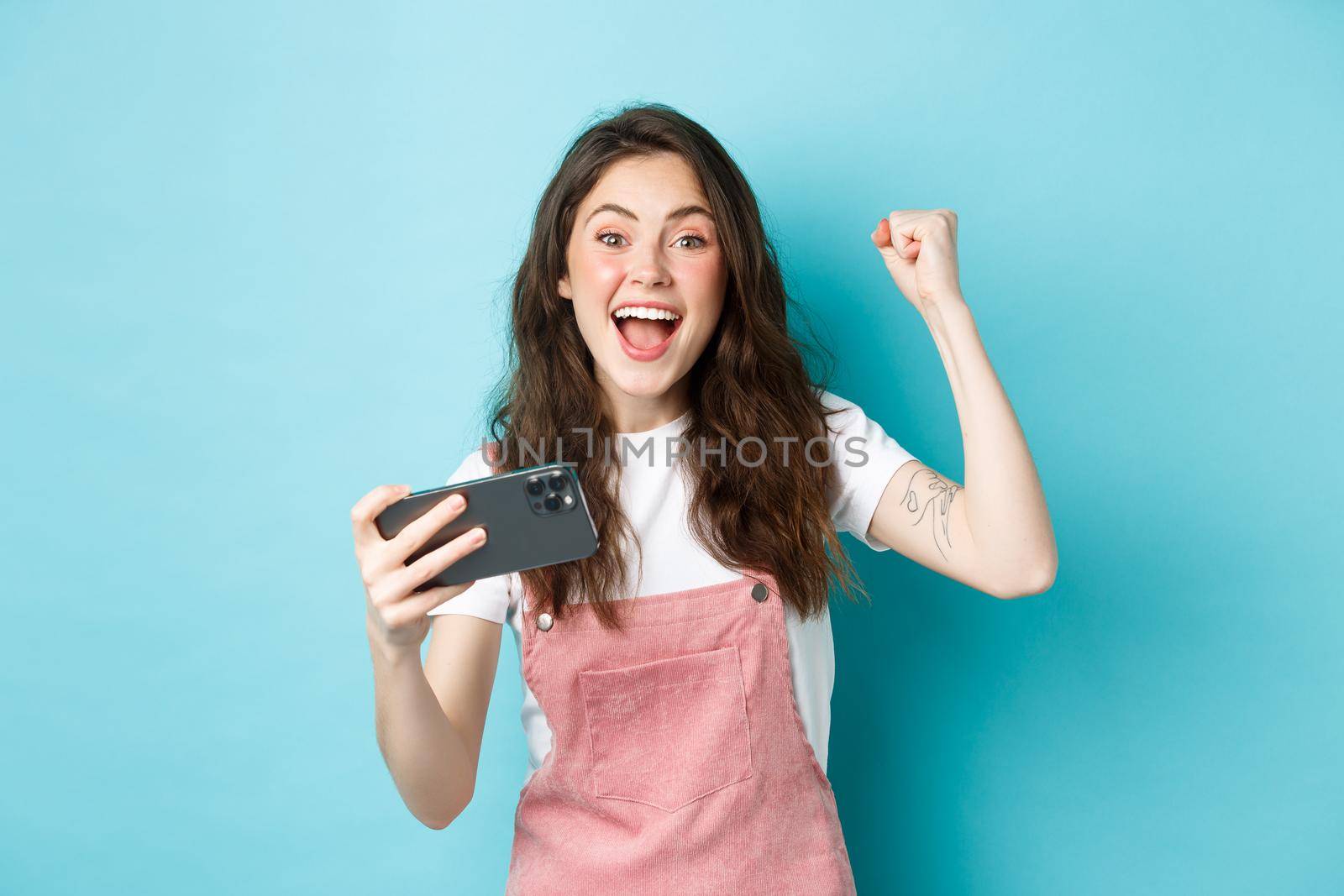 Excited beautful girl winning on mobile phone, holding smartphone and shouting yes with joyful face and fist pump, smiling amazed at camera, blue background by Benzoix