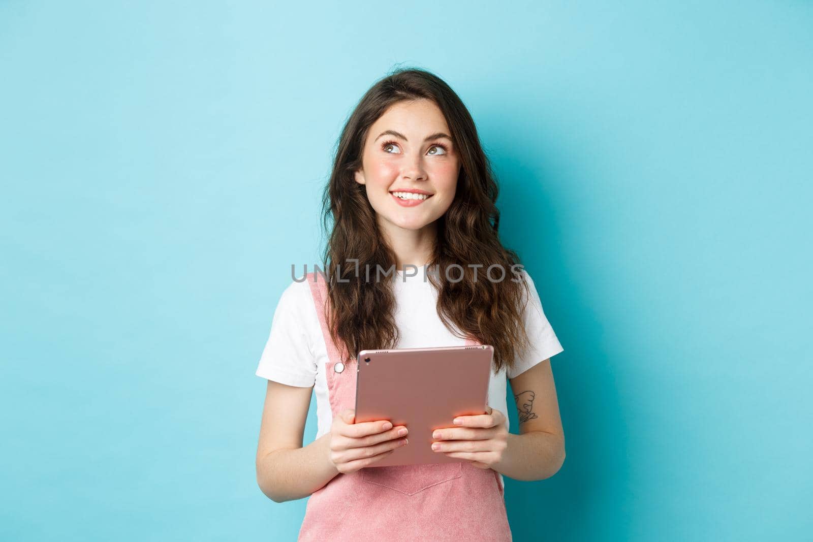 Beautiful dreamy girl smiling, holding tablet in hands and looking at upper left corner logo with thoughtful face, standing over blue background.