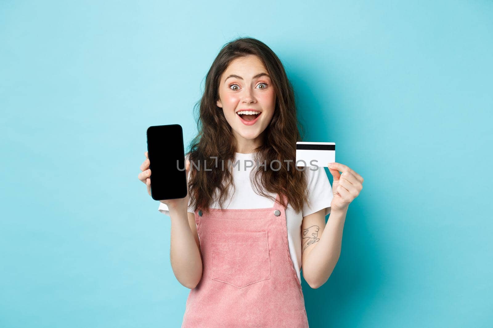 Look here. Excited young cute girl showing empty mobile screen and plastic credit card, look amazed at camera, standing over blue background.