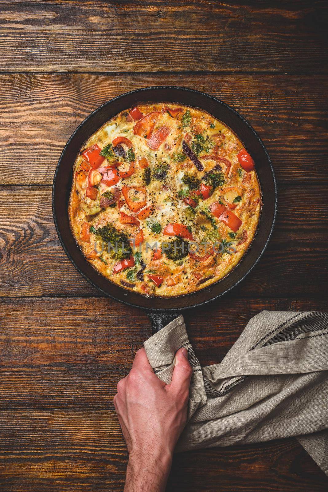 Vegetable frittata with broccoli, red bell pepper and herbs. Hand holds cast iron skillet