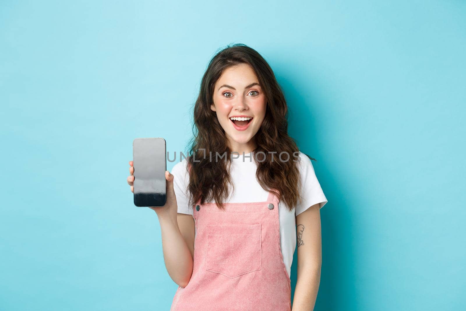Image of beautiful young woman look surprised and excited, showing empty smartphone screen, your logo or app on mobile display, standing over blue background.