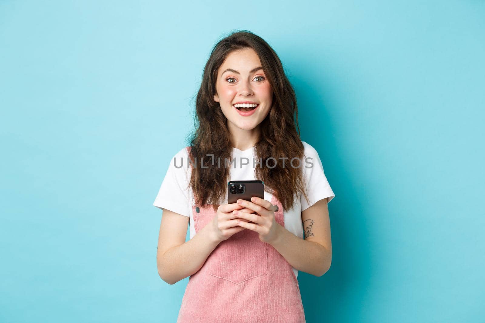 Portrait of excited brunette girl looking amazed at camera while using smartphone, holding mobile phone and smiling cheerful, standing against blue background by Benzoix