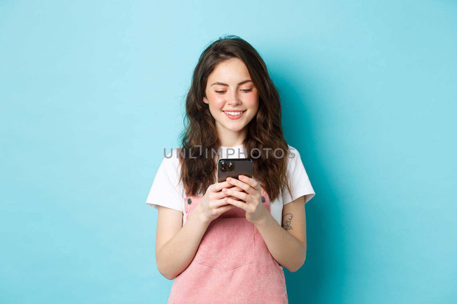 Portrait of young woman using smartphone, reading message or chatting in app, standing over blue background and smiling happy.