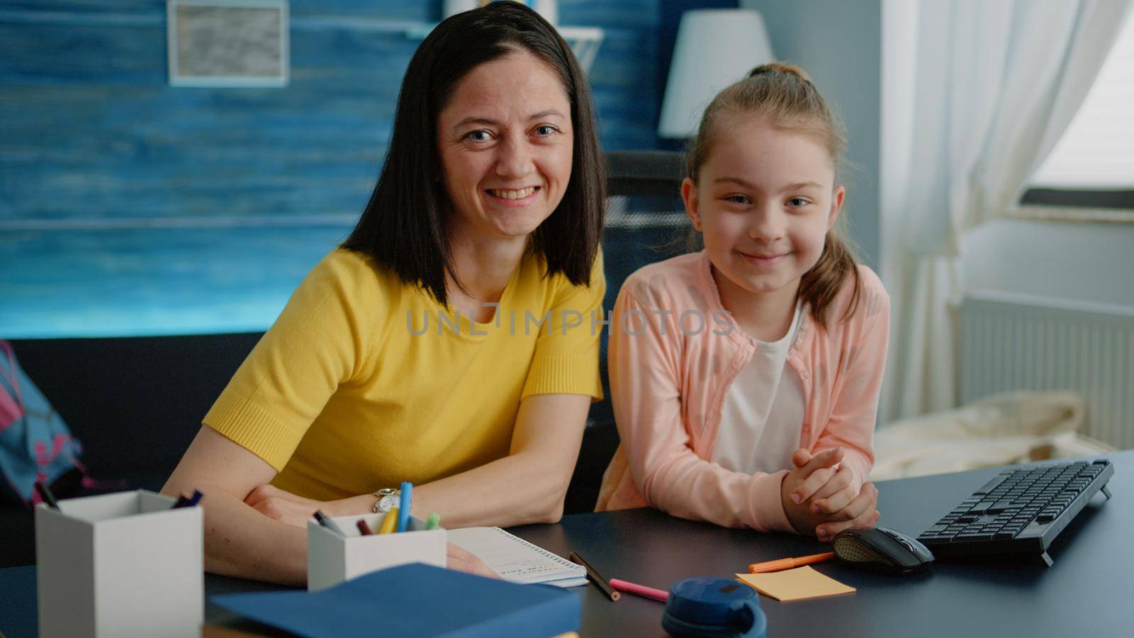 Affectionate mother sitting at desk with daughter for homework. Parent and little girl smiling and looking at camera, preparing for online remote class lesson at home. Adult and pupil