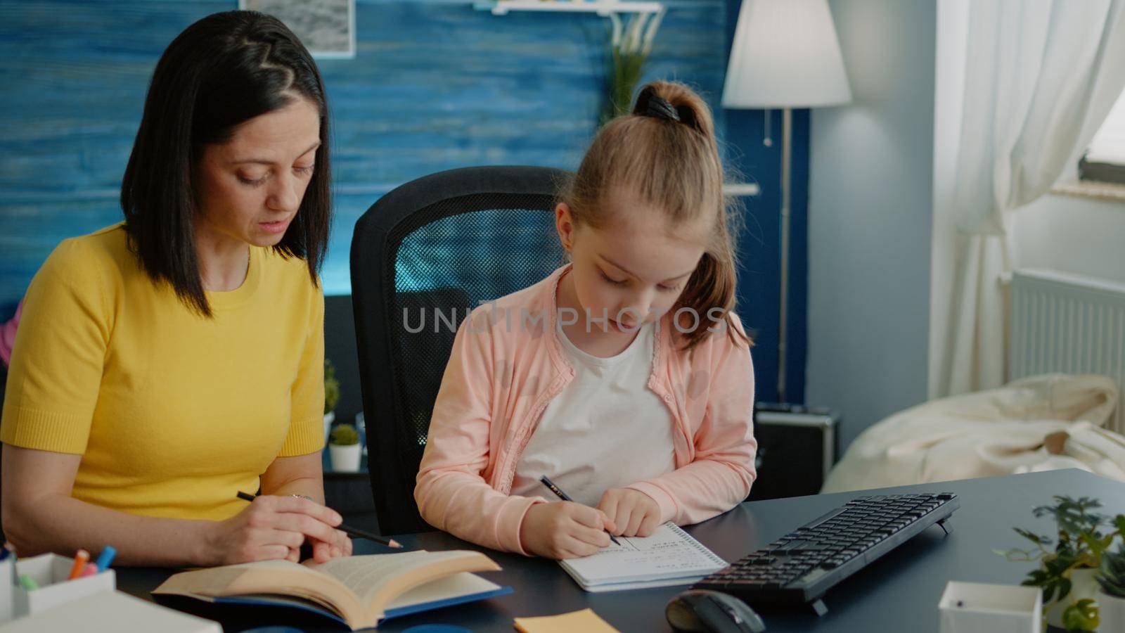 Parent using books to help little girl with homework at desk. Mother and young child doing tasks on notebook for online remote lesson. Pupil and adult learning for distance education