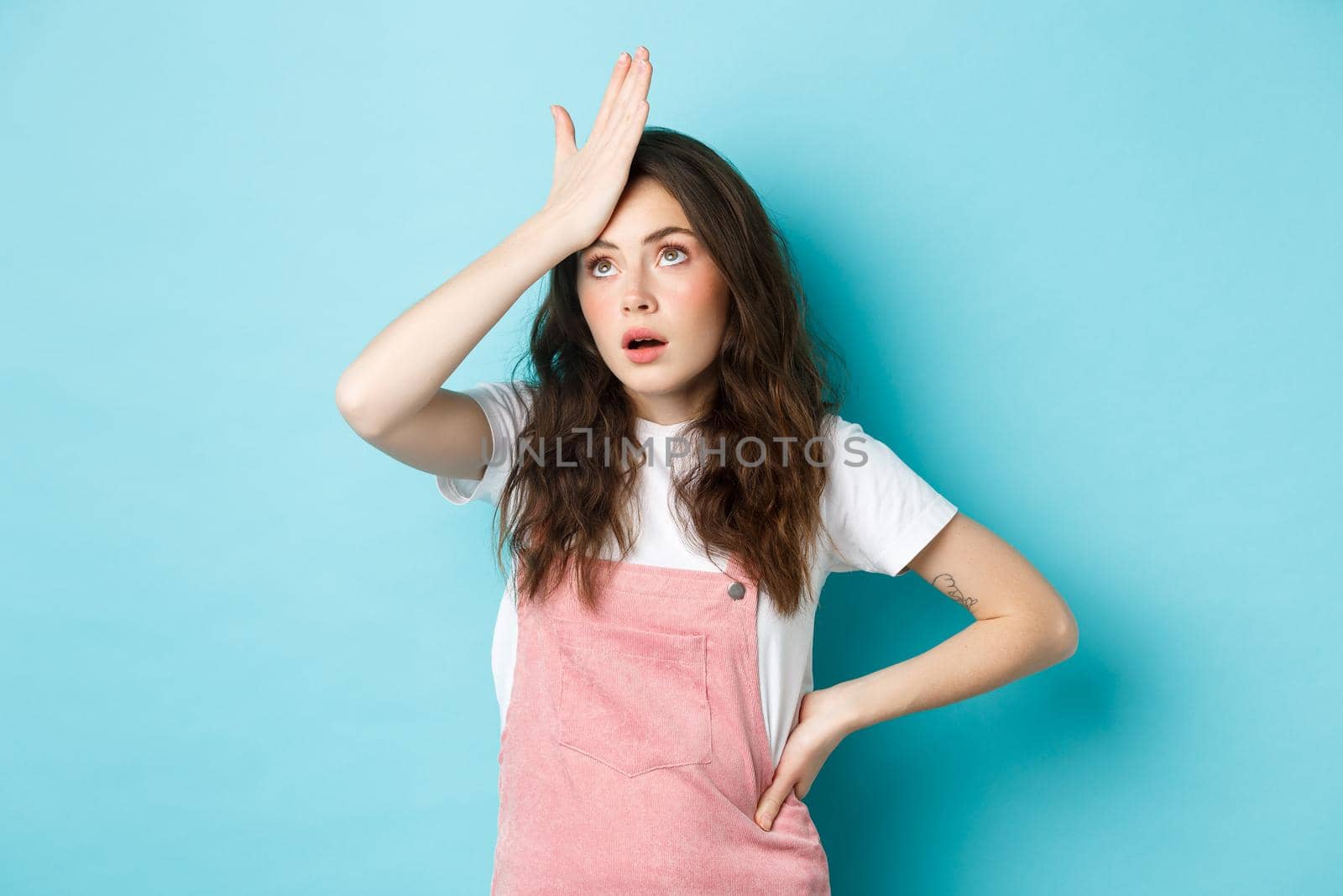 Annoyed and tired young woman in summer outfit, slap forehead and roll eyes, standing over blue background.