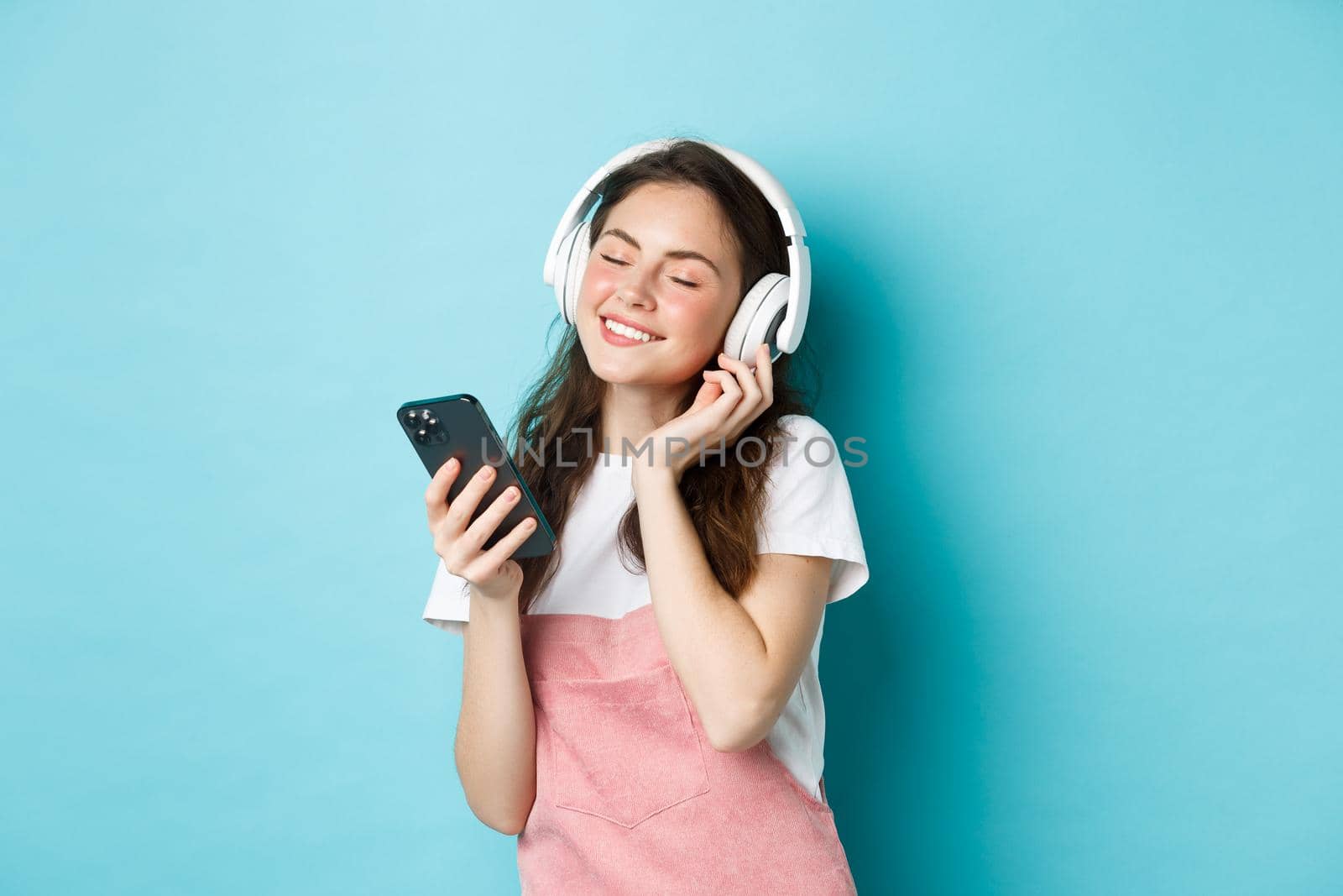 Beautiful woman enjoying song in headphones, close eyes and smile while listening music in headphones, holding smartphone in hand, standing over blue background by Benzoix