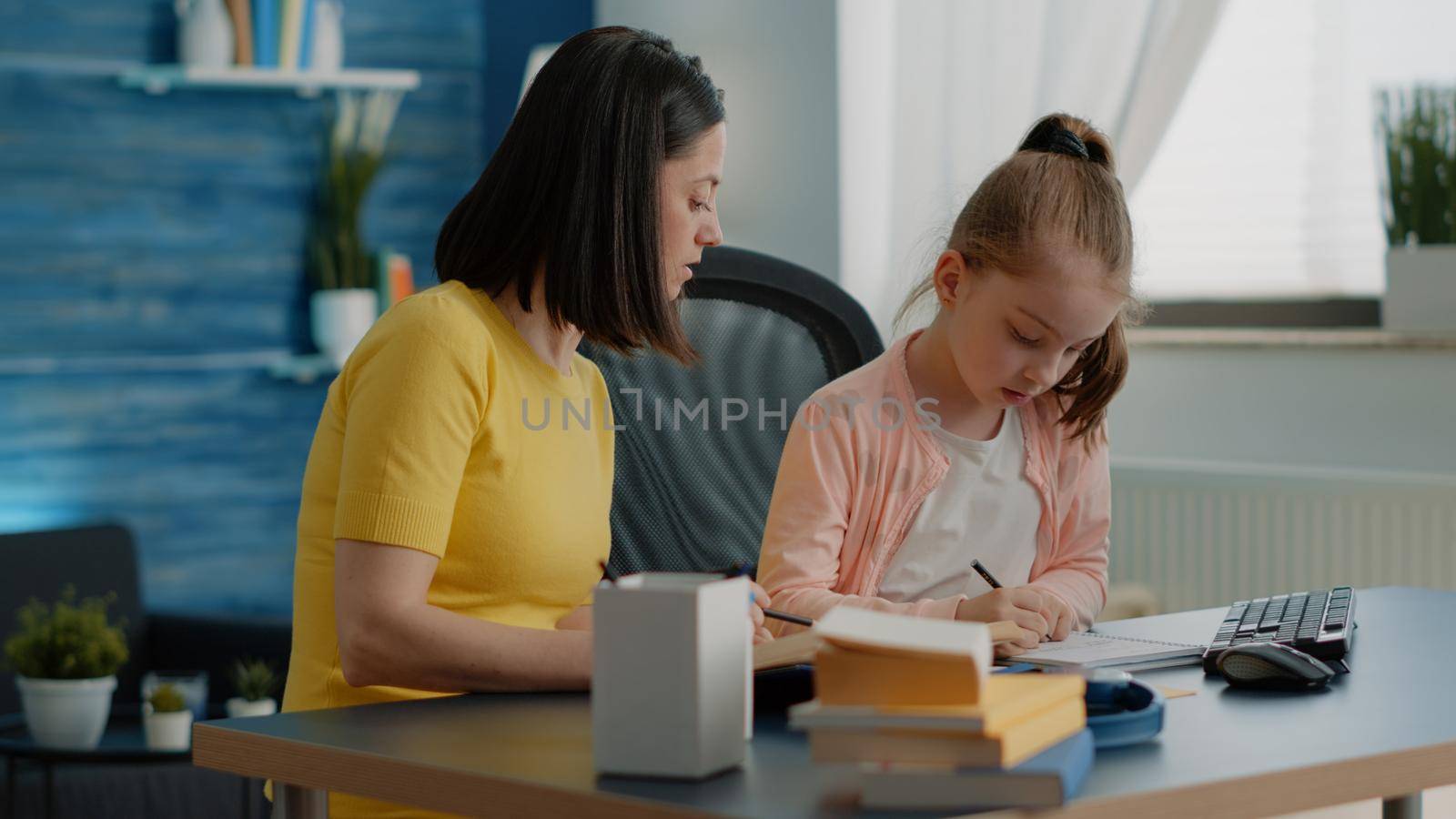 Pupil and mother doing homework together with books at desk. Young girl writing on textbook, preparing for online classes and school tasks with help from parent with supplies and book