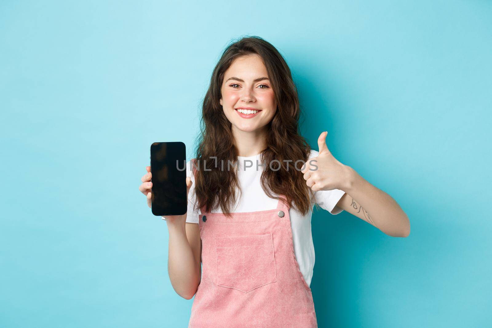 This is good. Smiling cheerful woman showng empty smartphone screen and thumb up to recommend app or online store, standing against blue background by Benzoix