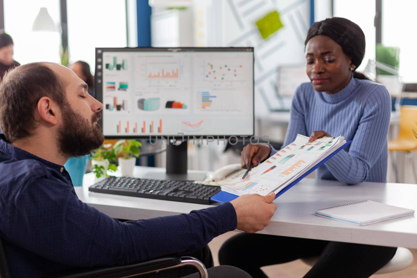 Black manager woman working with financial documents checking graphs talking with paralysed team leader with disabilities, reading raports sitting in wheelchair in start-up business office.