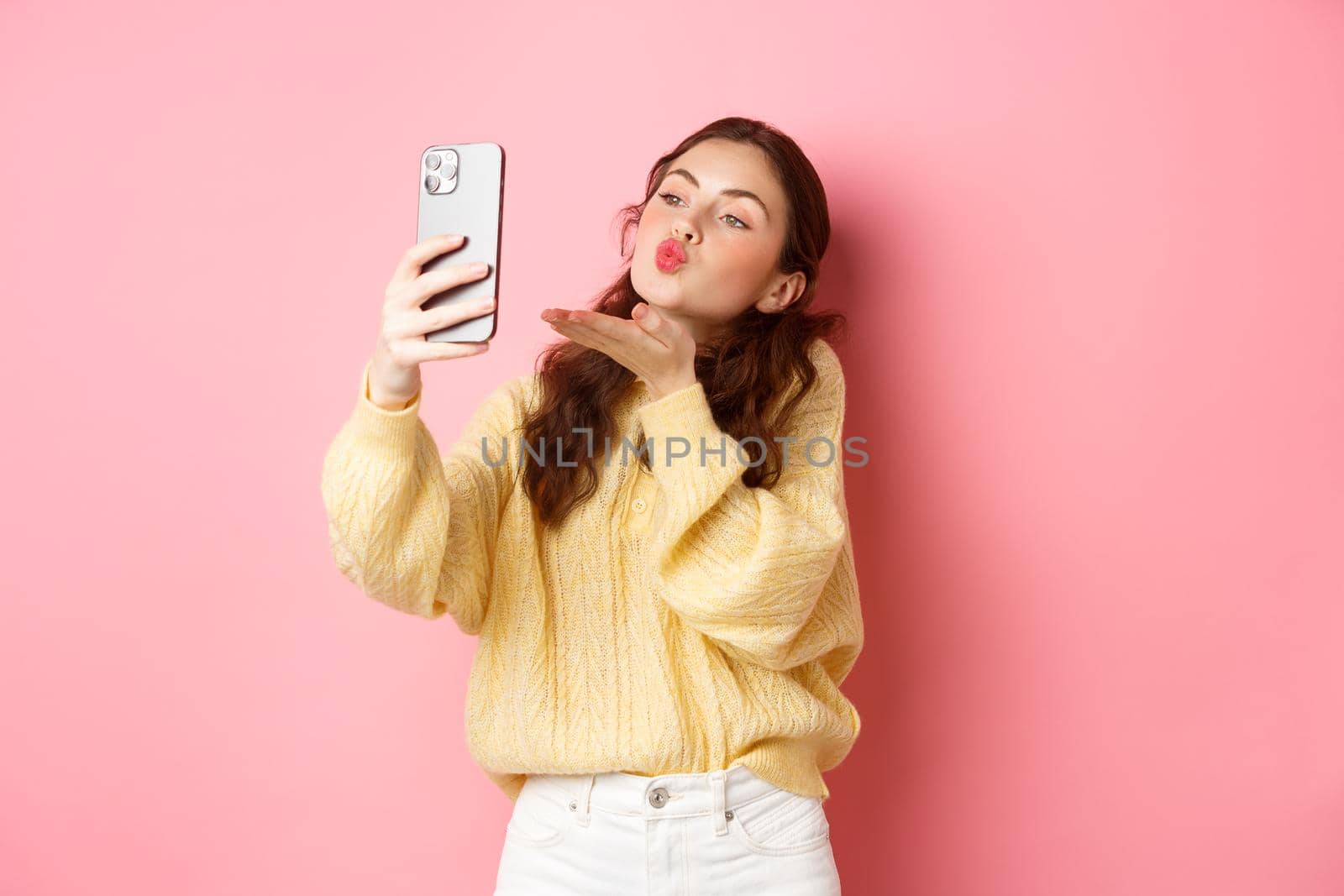 Beautiful flirty girl taking selfie on smartphone, sending air kiss during video chat, standing against pink background by Benzoix