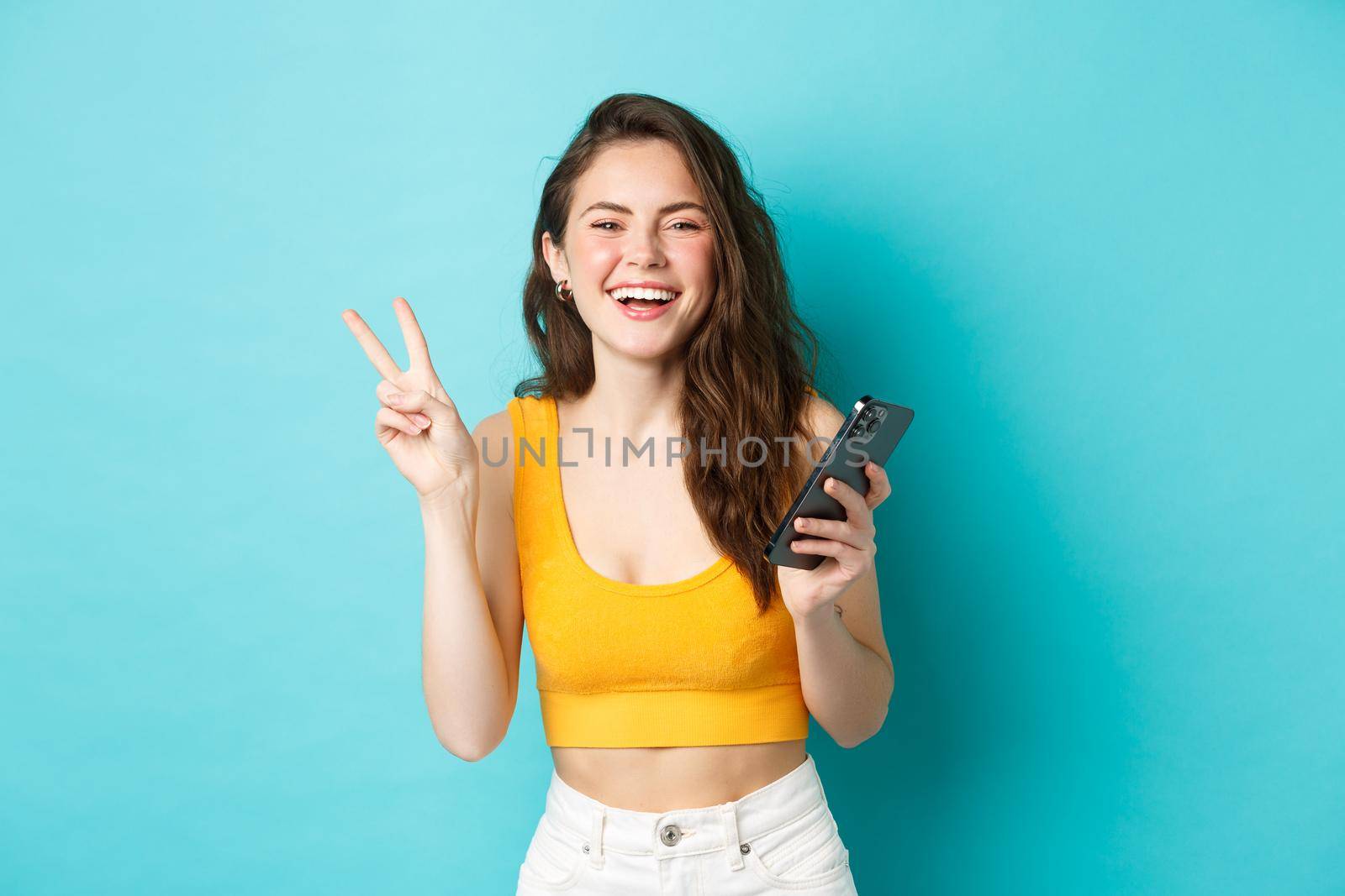 Technology and lifestyle concept. Happy attractive woman laughing, showing v-sign and using smartphone, chatting on mobile phone, standing over blue background.