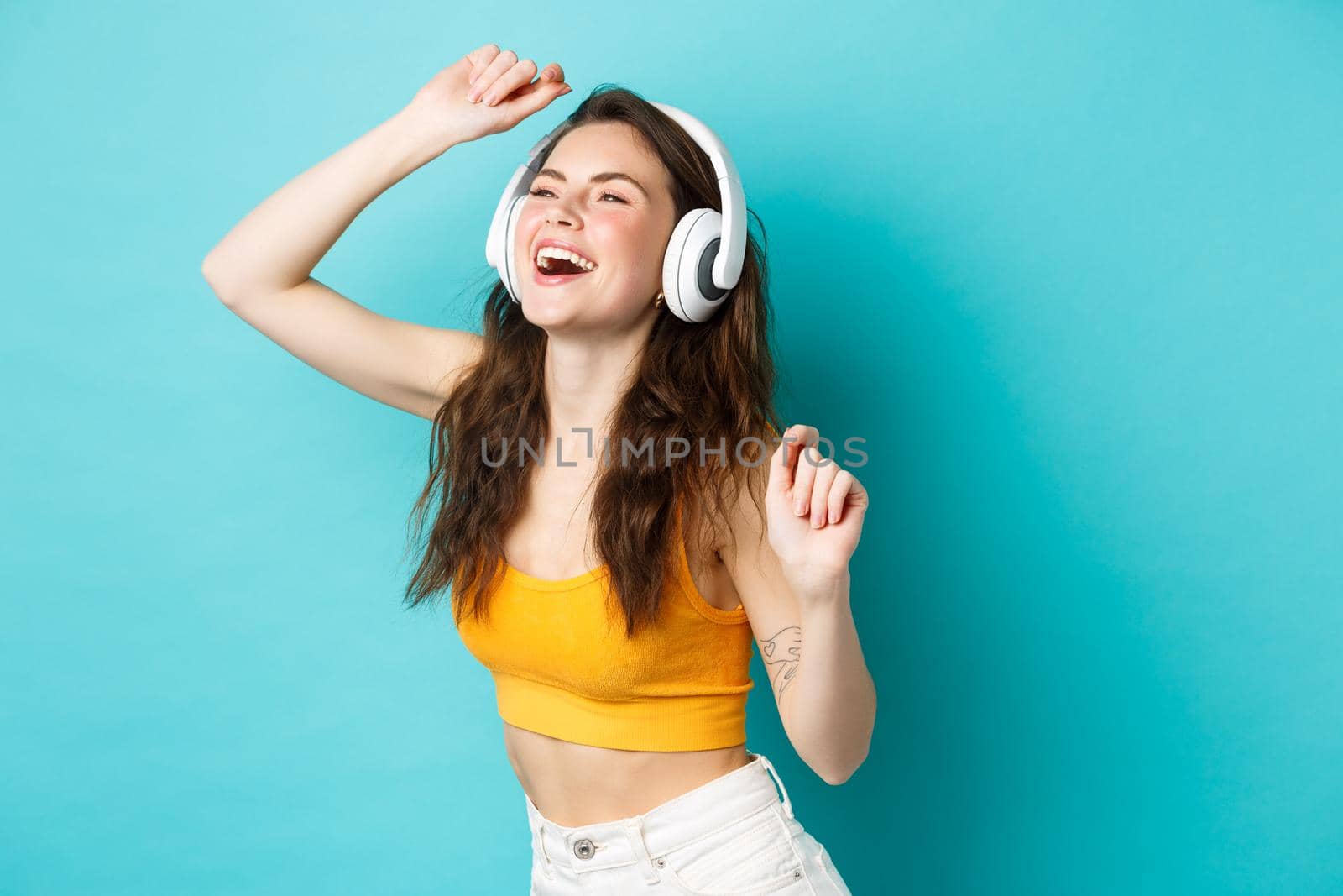 Beautiful summer girl dancing and laughing in headphones, listening music with carefree smile, standing over blue background.