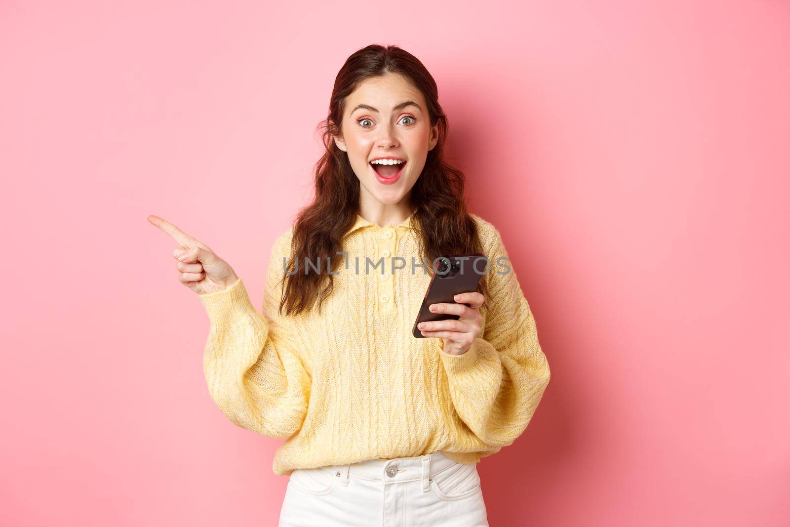 Excited young woman holding mobile phone, pointing finger aside at left copyspace, showing advertisement, found interesting promo online in smartphone, standing against pink background by Benzoix