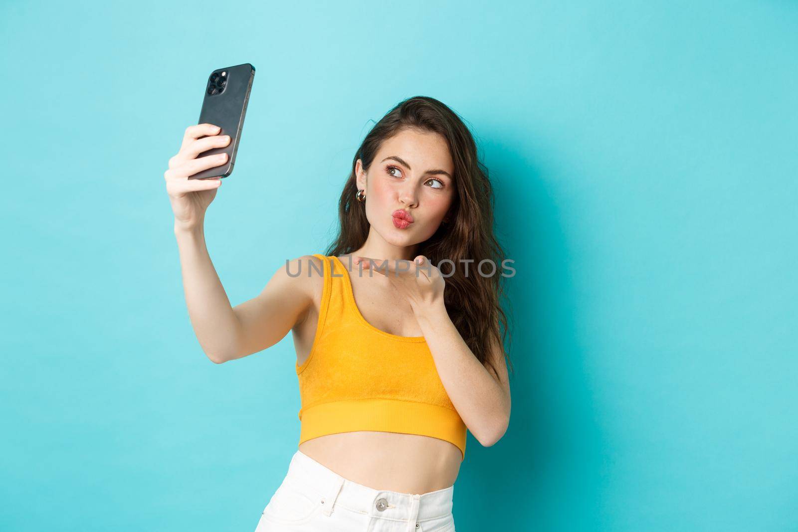 Technology and lifestyle concept. Stylish beautiful woman posing for selfie, sending air kiss at camera, using photo filters app on mobile phone, standing against blue background by Benzoix