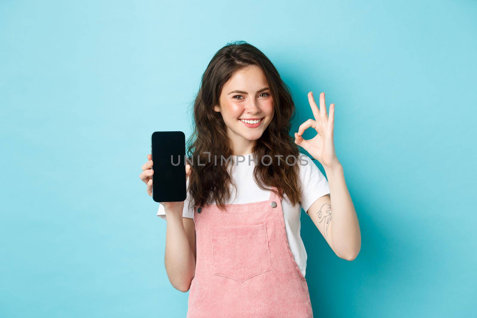 Portrait of modern stylish girl recommending online shop or mobile app, showing okay sign with empty smartphone screen, nod in approval, smiling satisfied, blue background.
