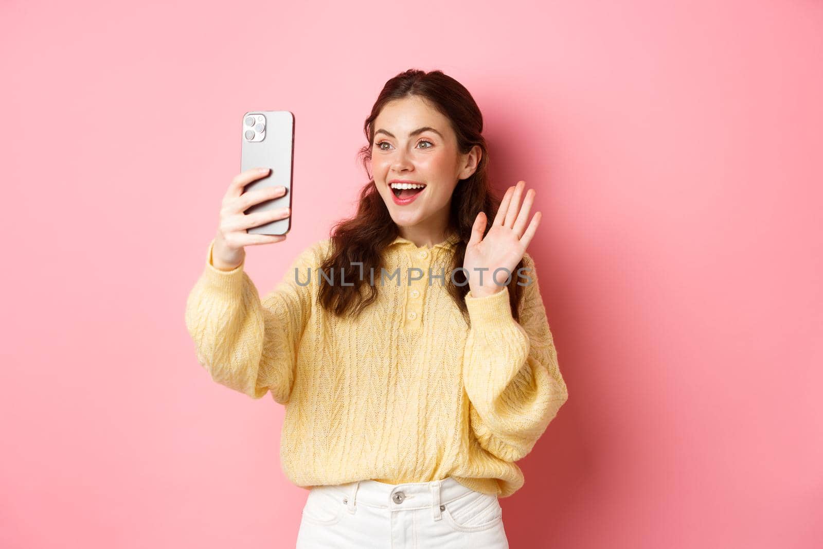 Cheerful girl video chatting on smartphone, waving hand at phone camera and smiling happy, say hello, standing against pink background.