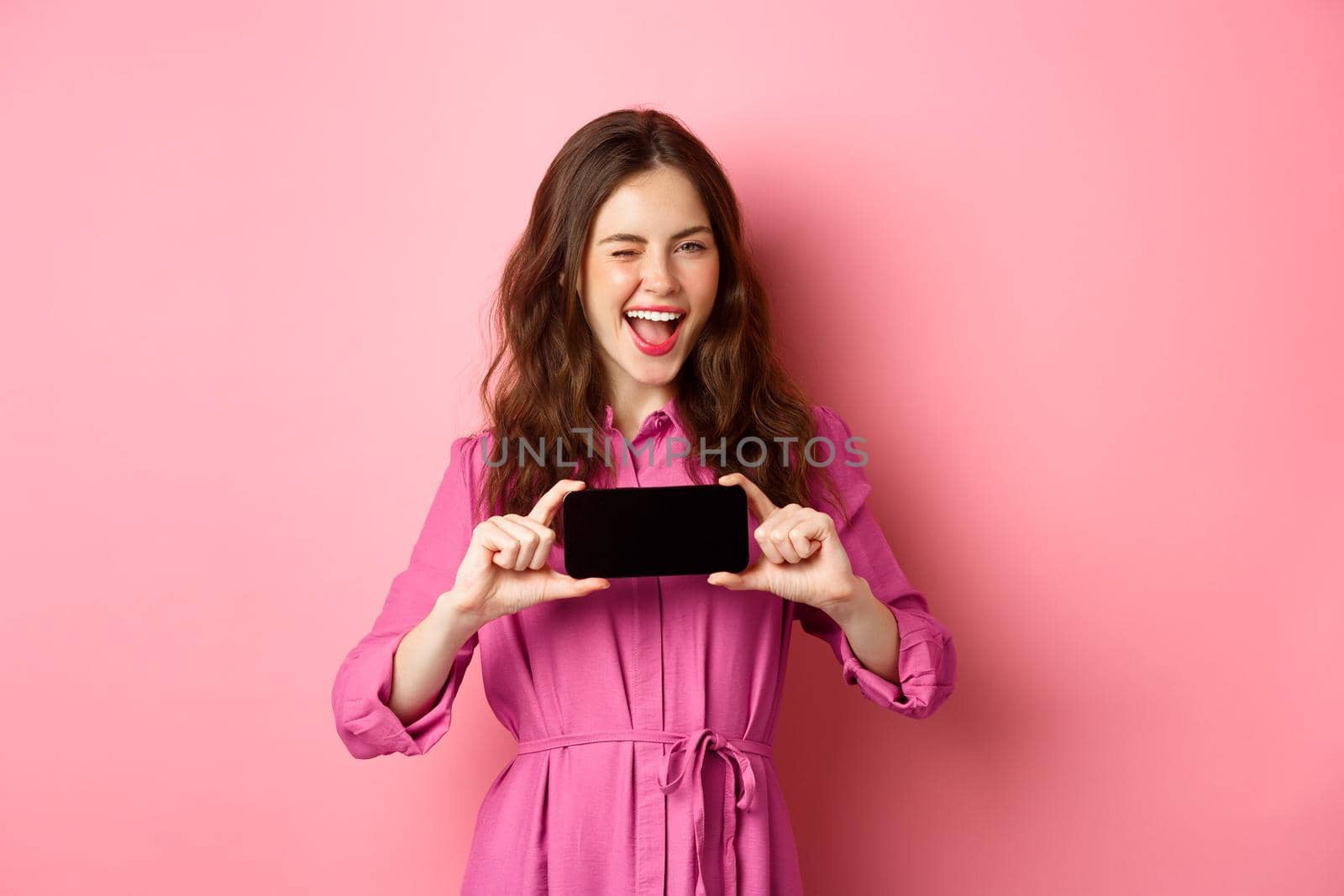 Technology concept. Cheerful lady in dress wink at you, smiling and showing mobile phone screen horizontally, standing over pink background.