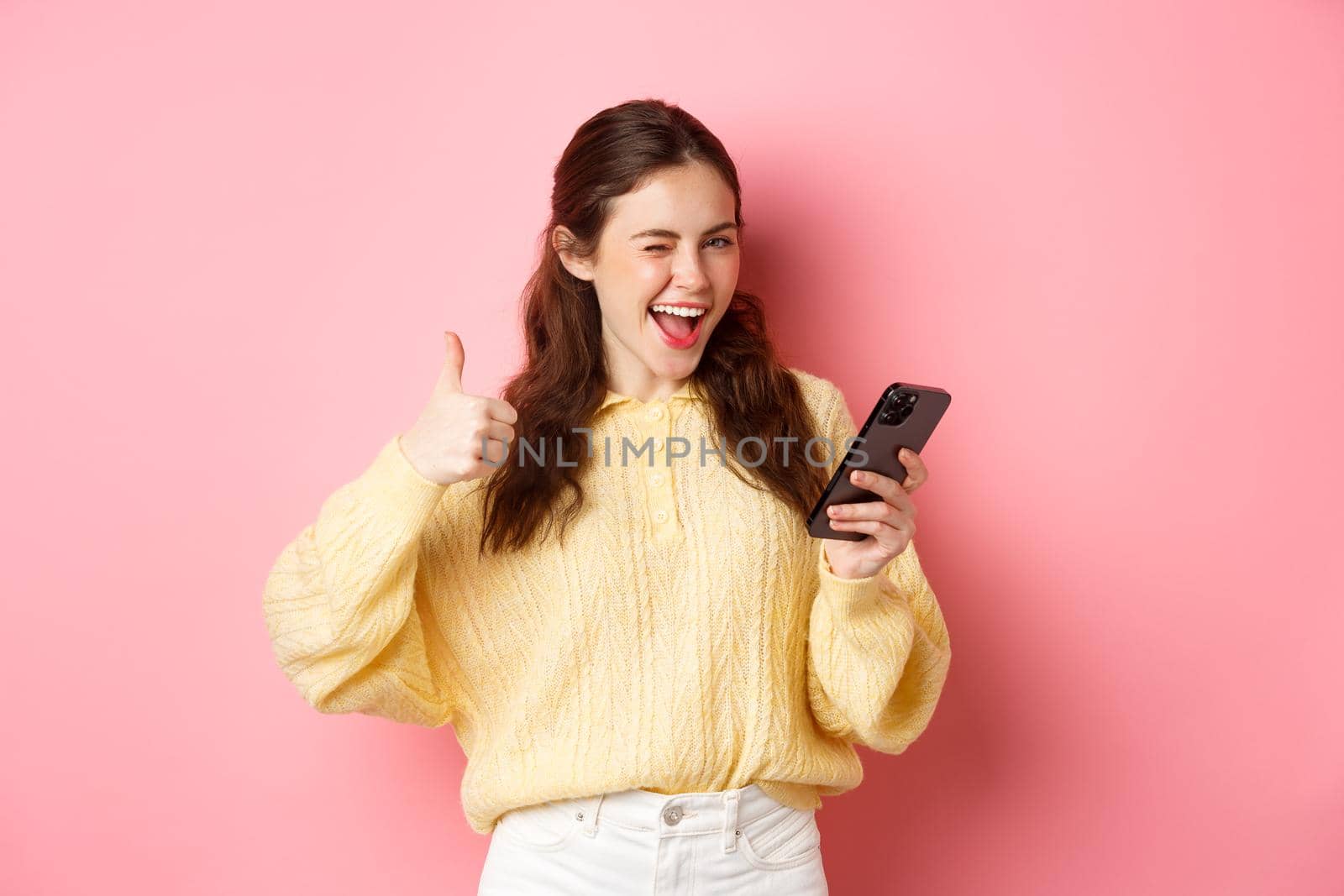 Happy pretty woman saying yes, holding mobile phone and making thumbs up gesture, approve good thing, standing against pink background.