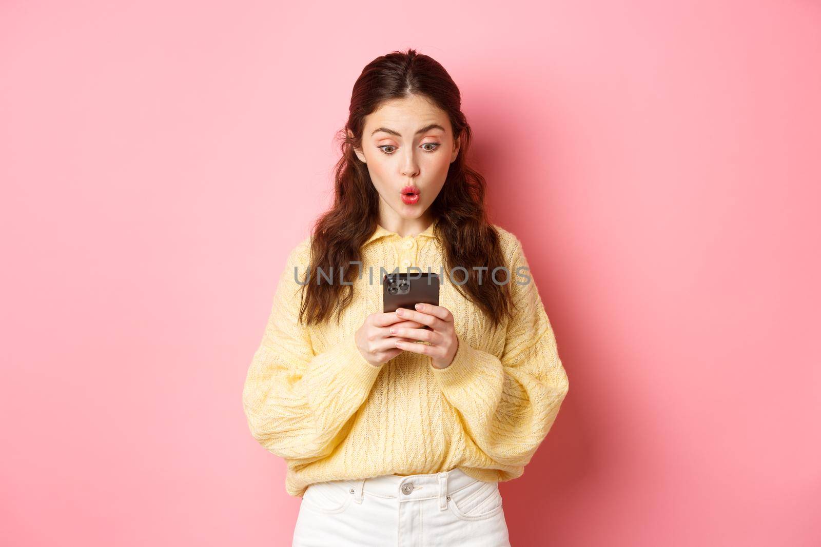 Portrait of surprised young woman reading message on smartphone and say wow, look curious at mobile screen, standing against pink background.