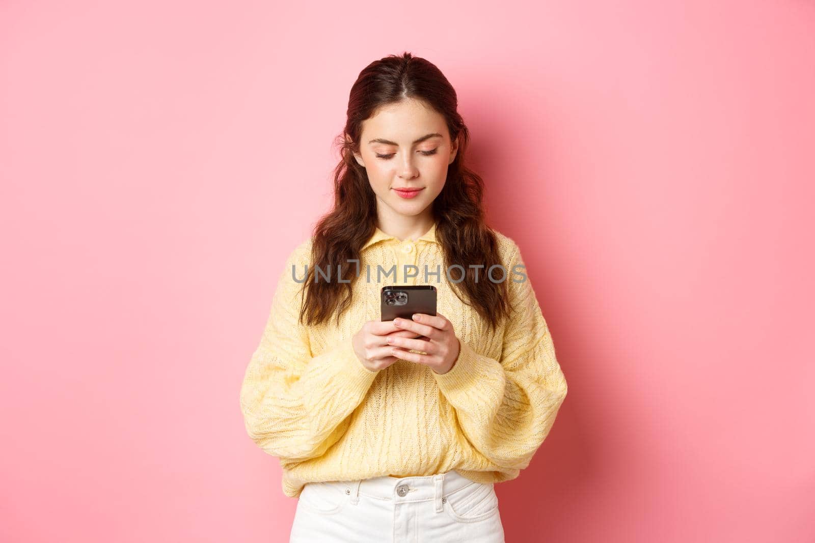 Technology. Young smiling woman chatting on social media, reading message on smartphone, standing against pink background.