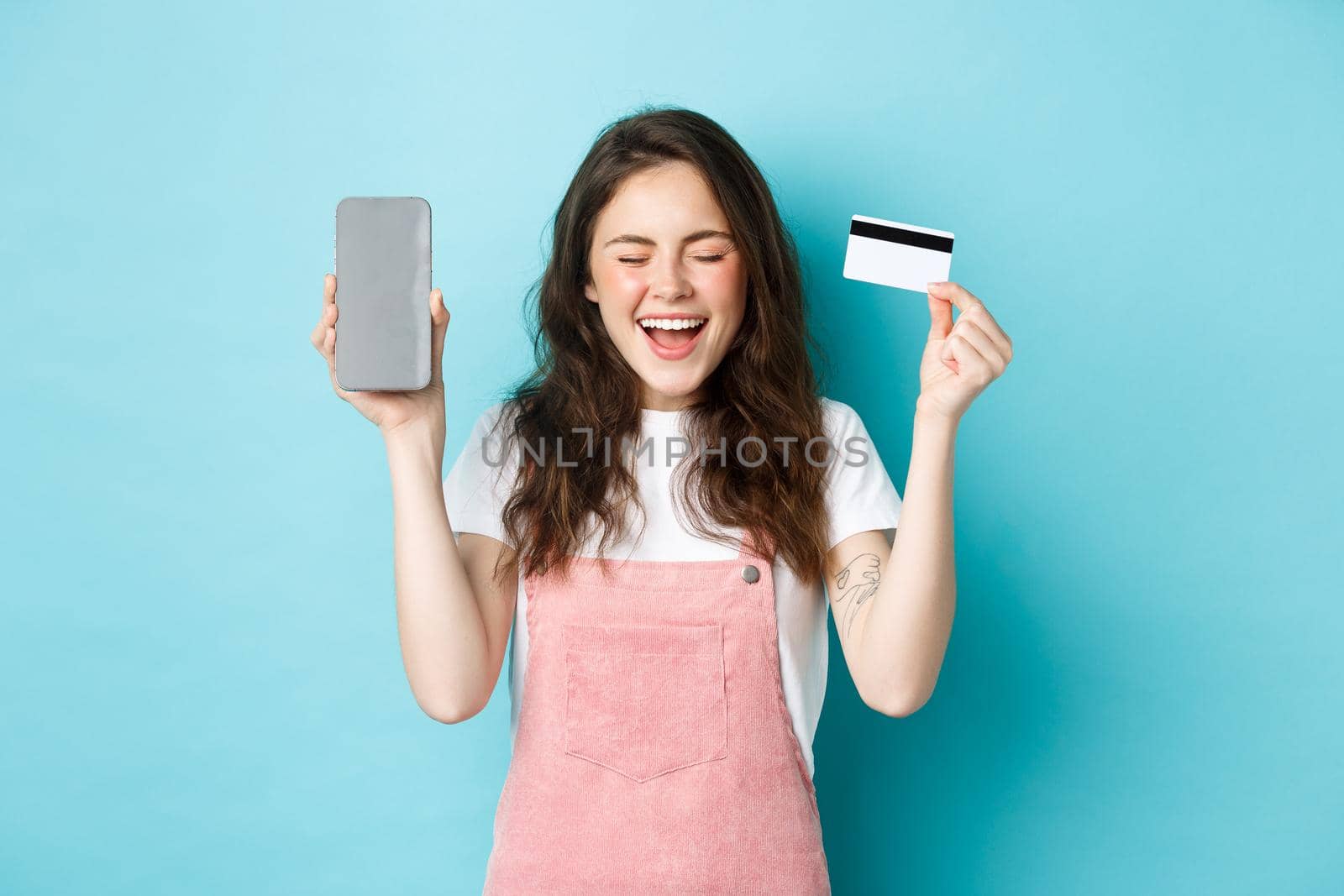 Excited attractive woman scream of joy, showing empty smartphone screen and plastic credit card, raising hands up and smiling happy, standing over blue background by Benzoix