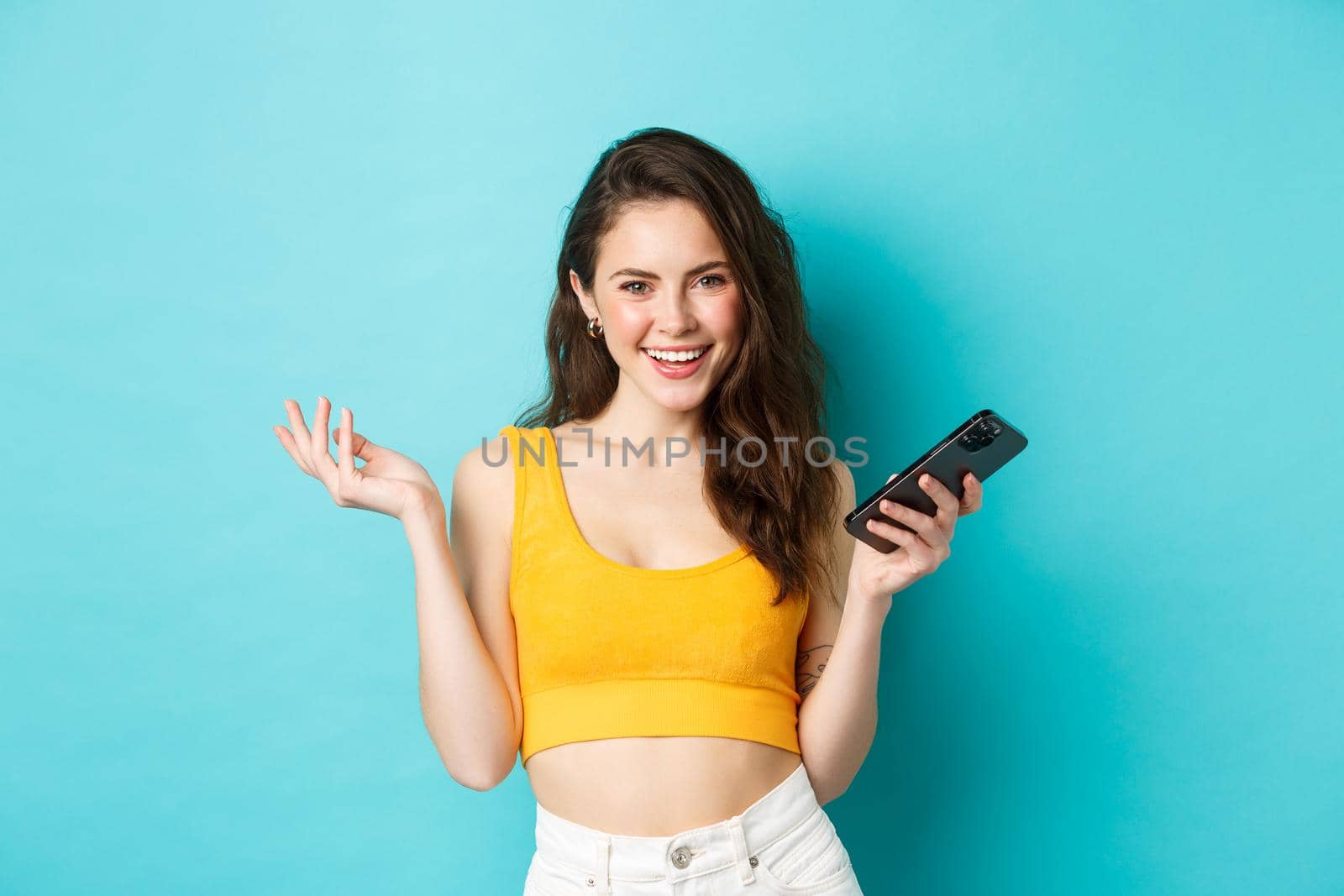 Stylish happy woman holding smartphone, laughing and smiling self-assured, chatting on mobile phone, standing over blue background.