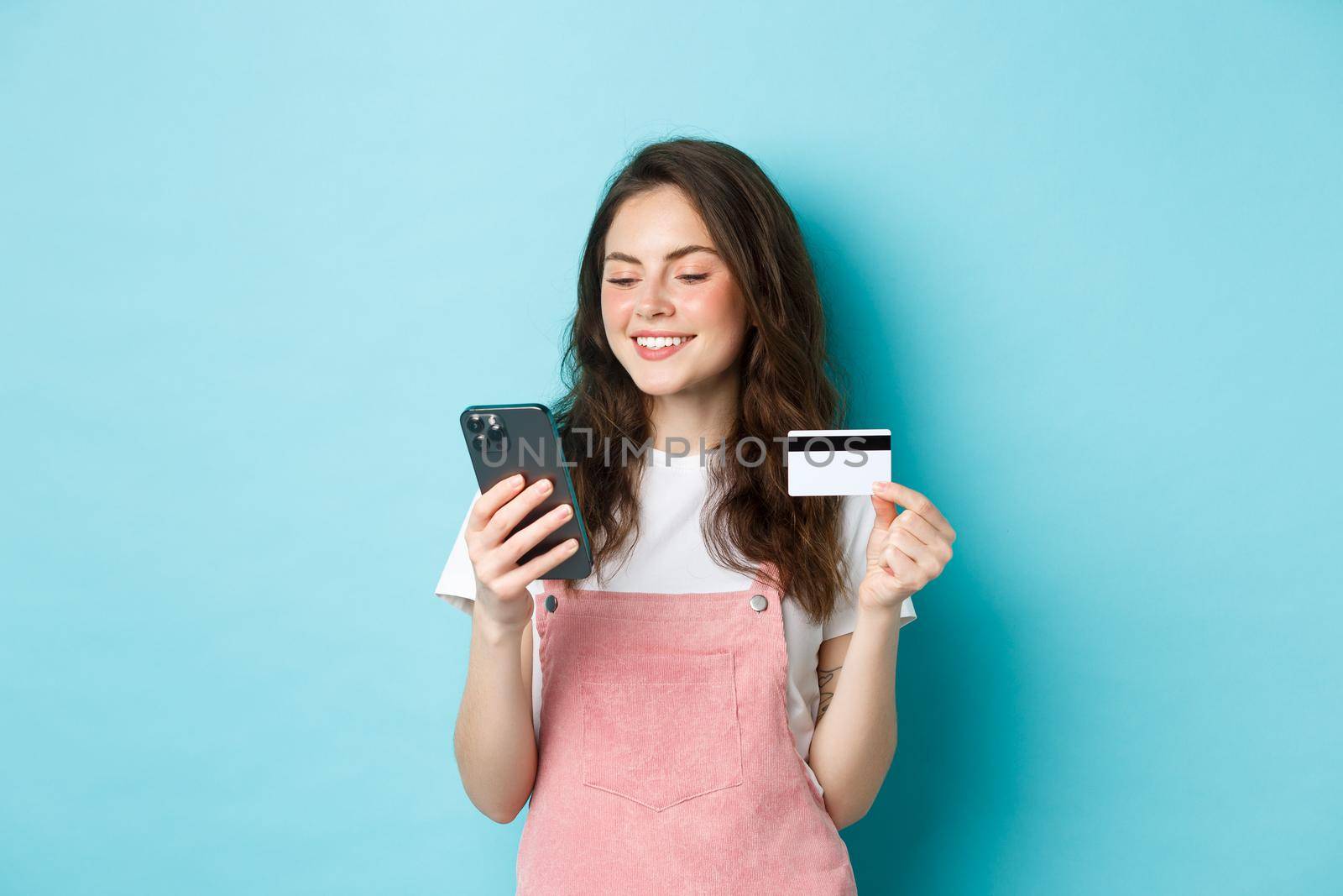 Young modern girl paying online with credit card, type info on smartphone, looking at screen with pleased face, shopping in app, standing over blue background by Benzoix