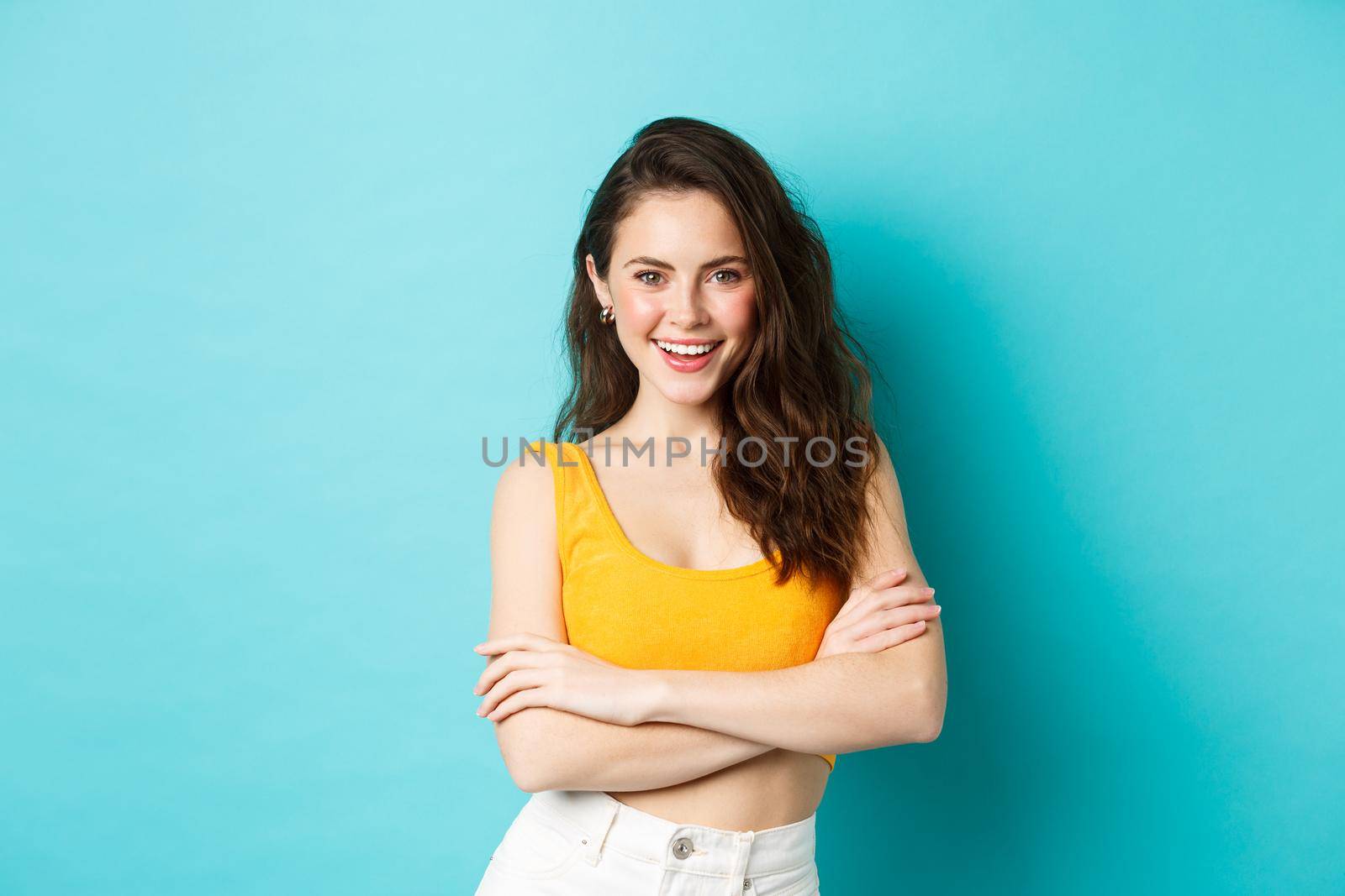 Summer and lifestyle concept. Confident and sassy attractive woman in cropped top looking determined at camera, cross arms on chest and smiling, standing against blue background.