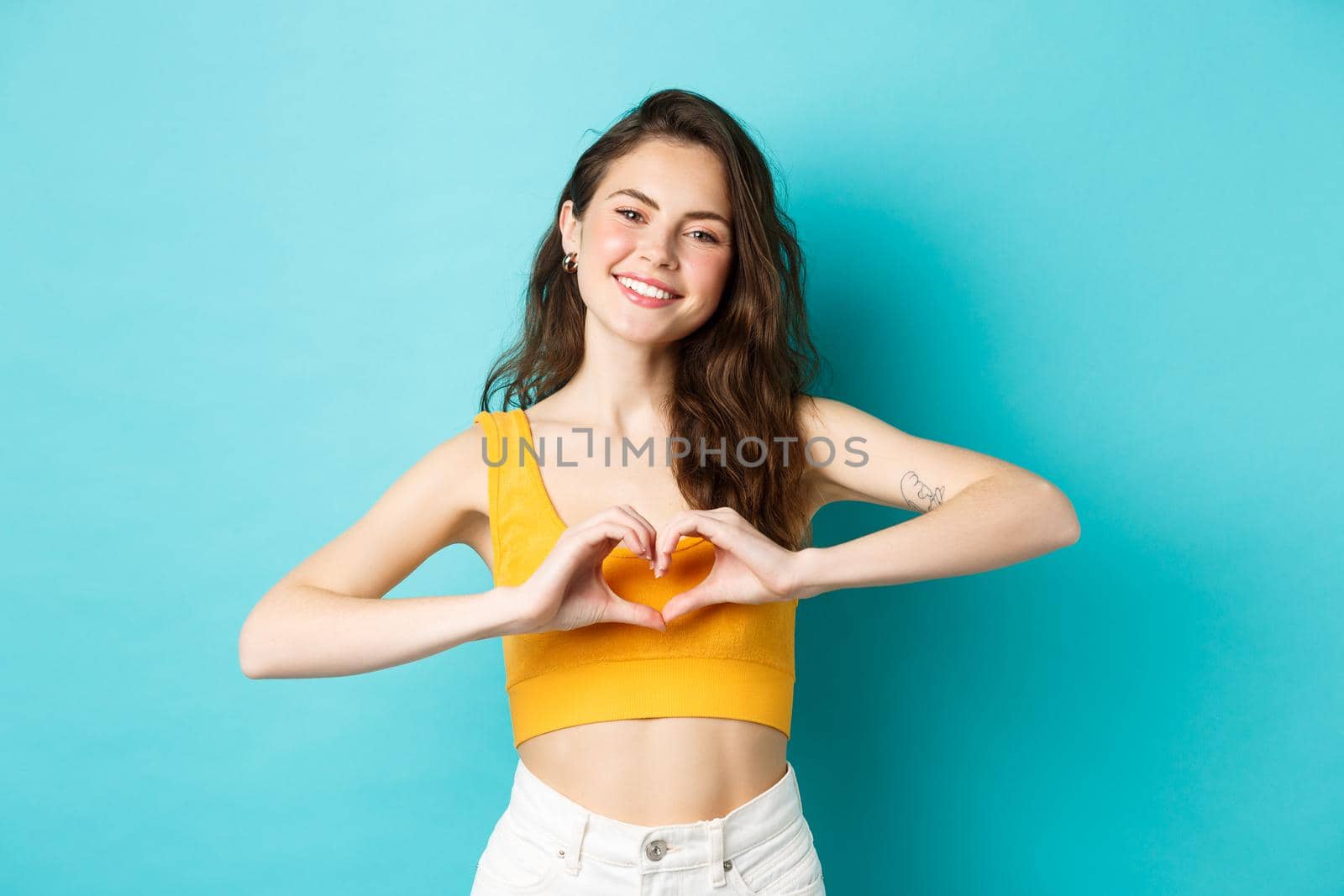 Summer holidays and emotions concept. Young romantic girl showing I love you heart gesture and smiling, standing over blue background.