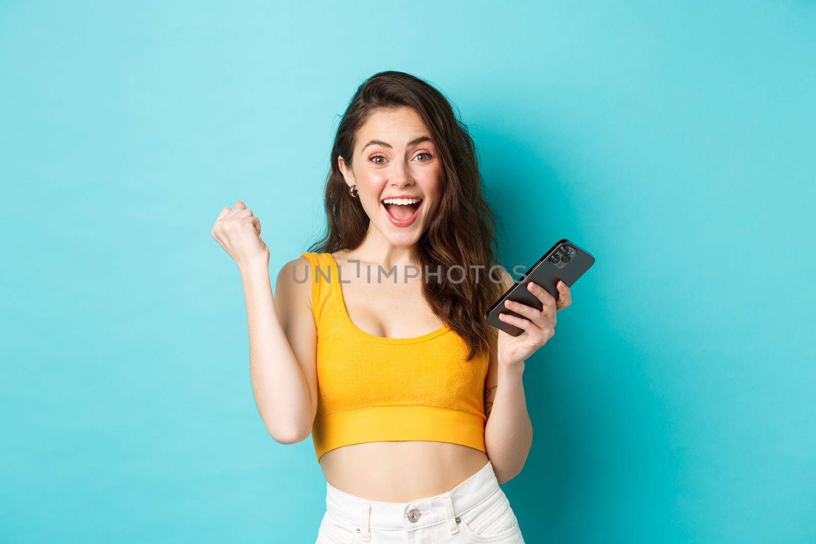 Technology and lifestyle concept. Excited woman winning online, holding smartphone and making yes fist pump gesture, celebrating, achieve goal in app, blue background by Benzoix