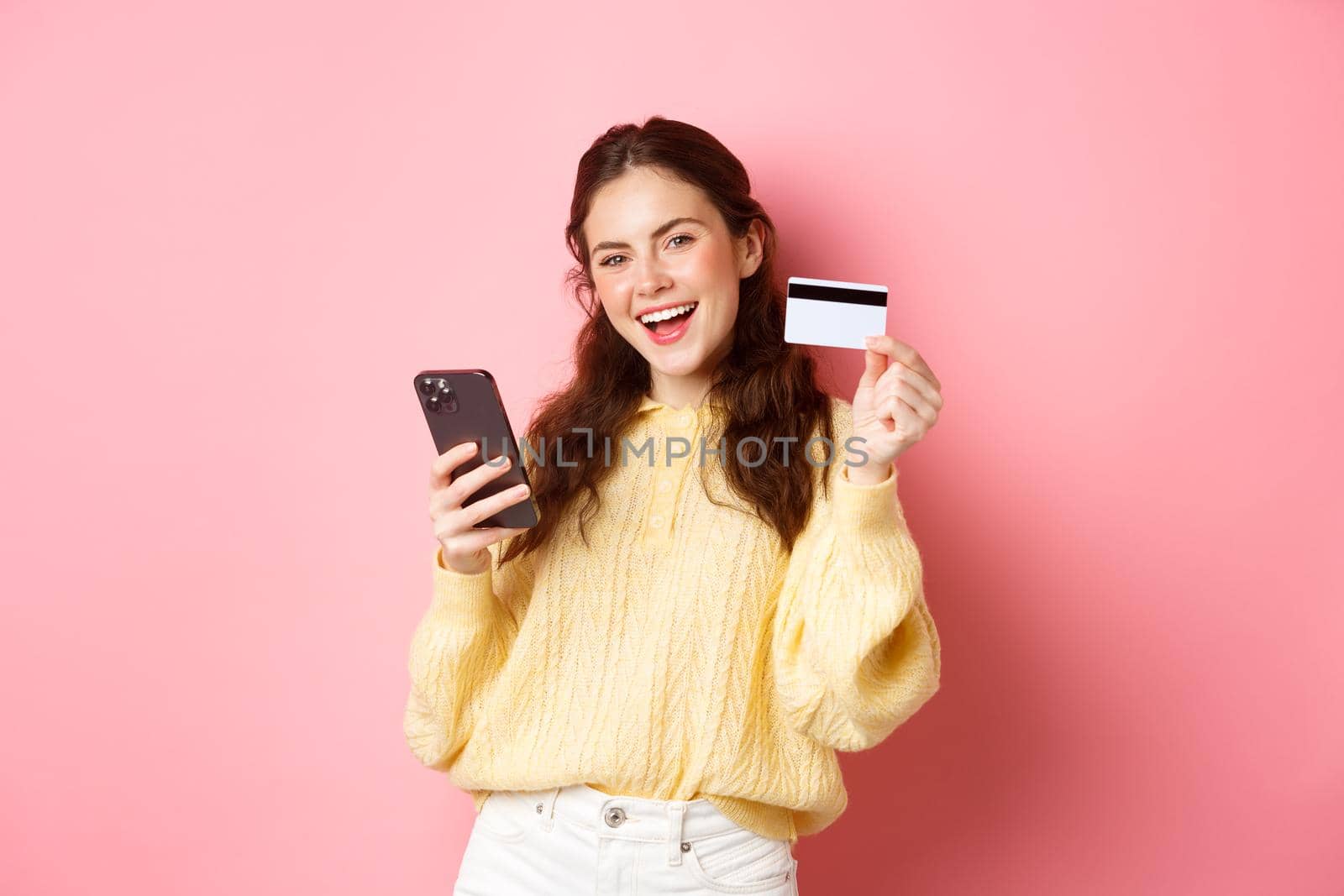Technology and online shopping. Happy and satisfied female client, showing plastic credit card and using mobile phone app to pay online, standing against pink background.