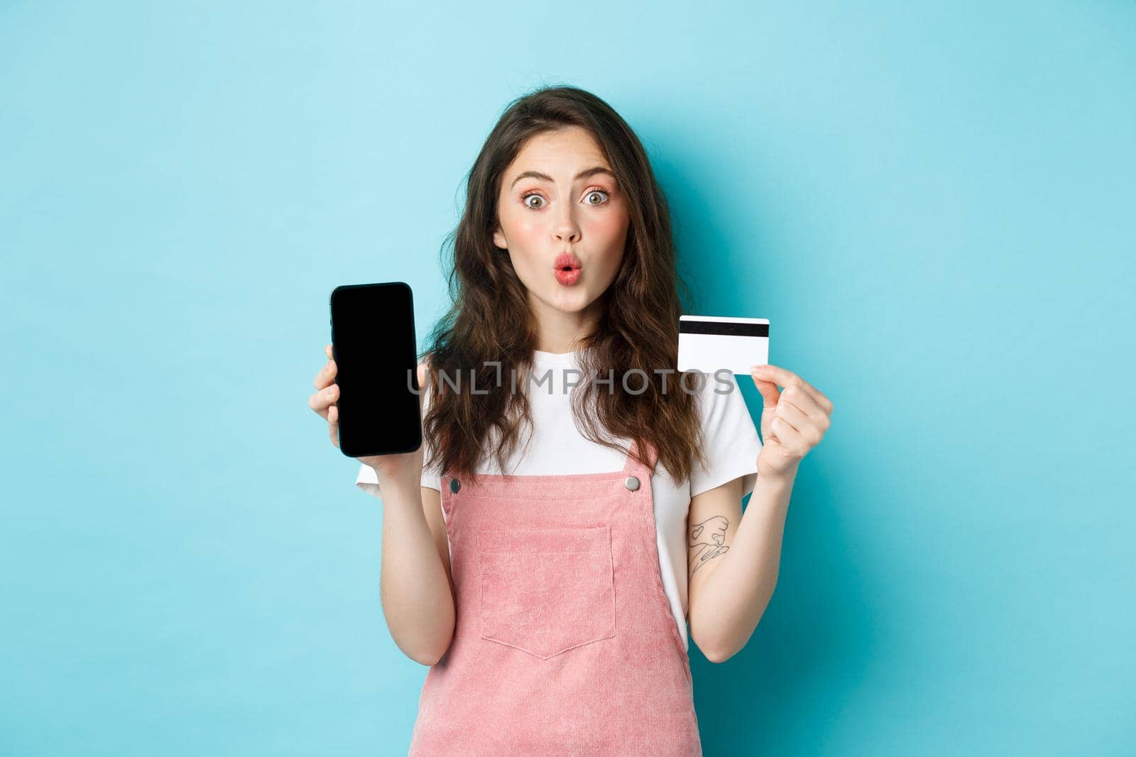 Portrait of young beautiful woman showing plastic credit card and empty smartphone screen, look amused and interested, showing interesting app, standing over blue background.