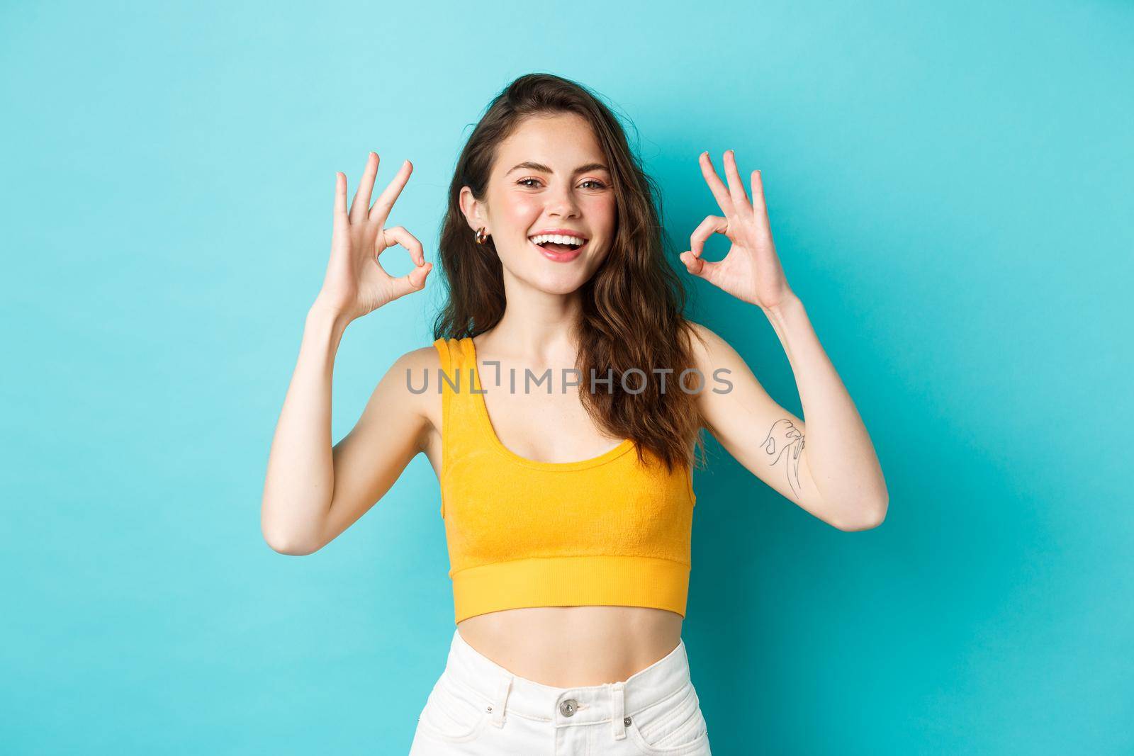Young carefree woman say yes and nod in approval, showing okay gestures to praise good choice, show perfect body in summer outfit, ready for vacation, standing over blue background.