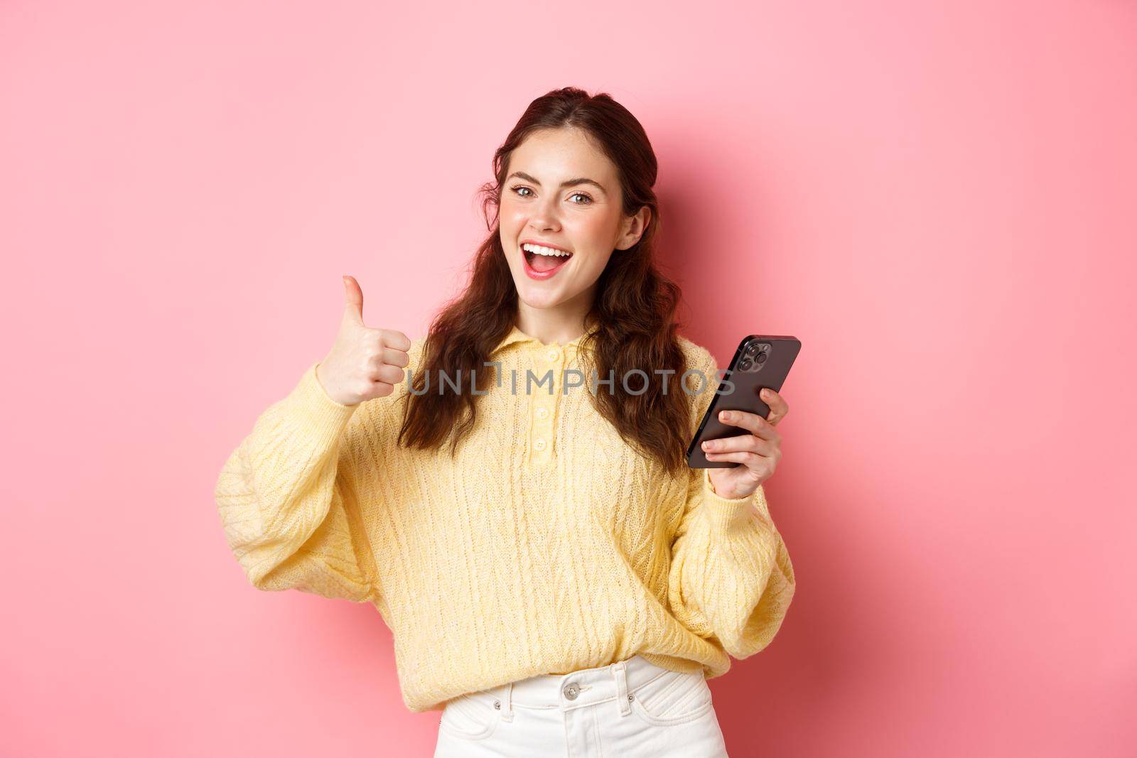 Portrait of attractive glamour girl showing thumbs up after using app on smartphone, online shopping application, standing against pink background, smiling satisfied.