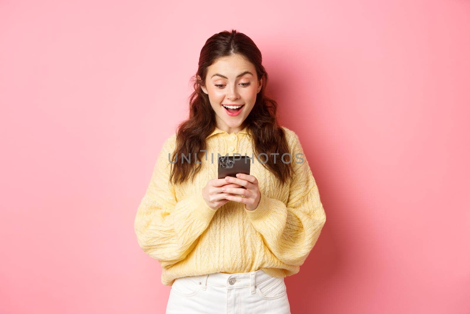 Portrait of excited brunette woman gasping amazed, reading message on smartphone, look surprised at mobile phone, standing over pink background.