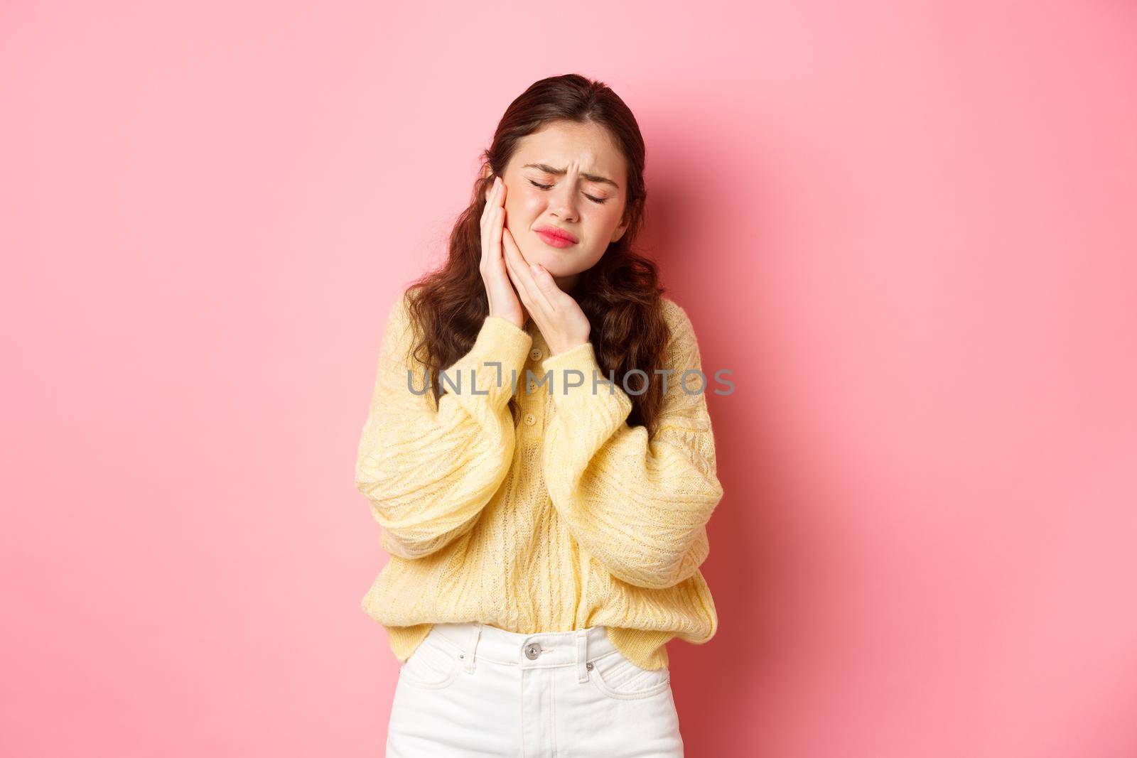 Young woman with terrible toothache, touching swallen cheek and grimacing from painful feeling, having tooth decay, need appointment to dentist, standing against pink background by Benzoix
